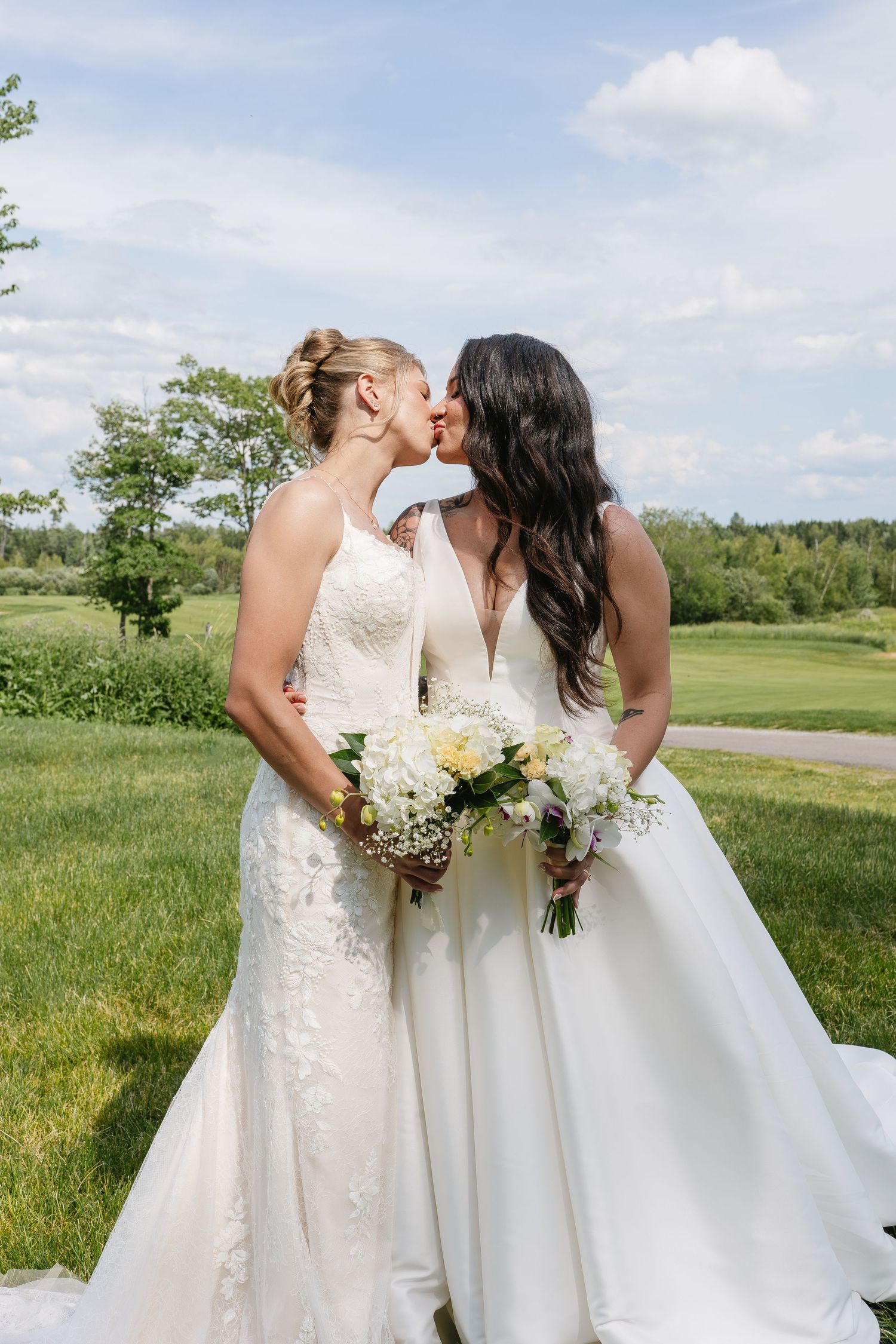 Two brides in white wedding gowns share a kiss while holding bouquets of white flowers outdoors.