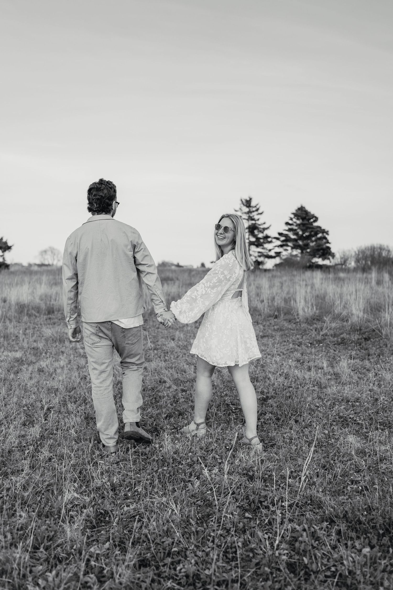 Black and white photo of a couple holding hands in a field, woman in white dress looks back smiling.