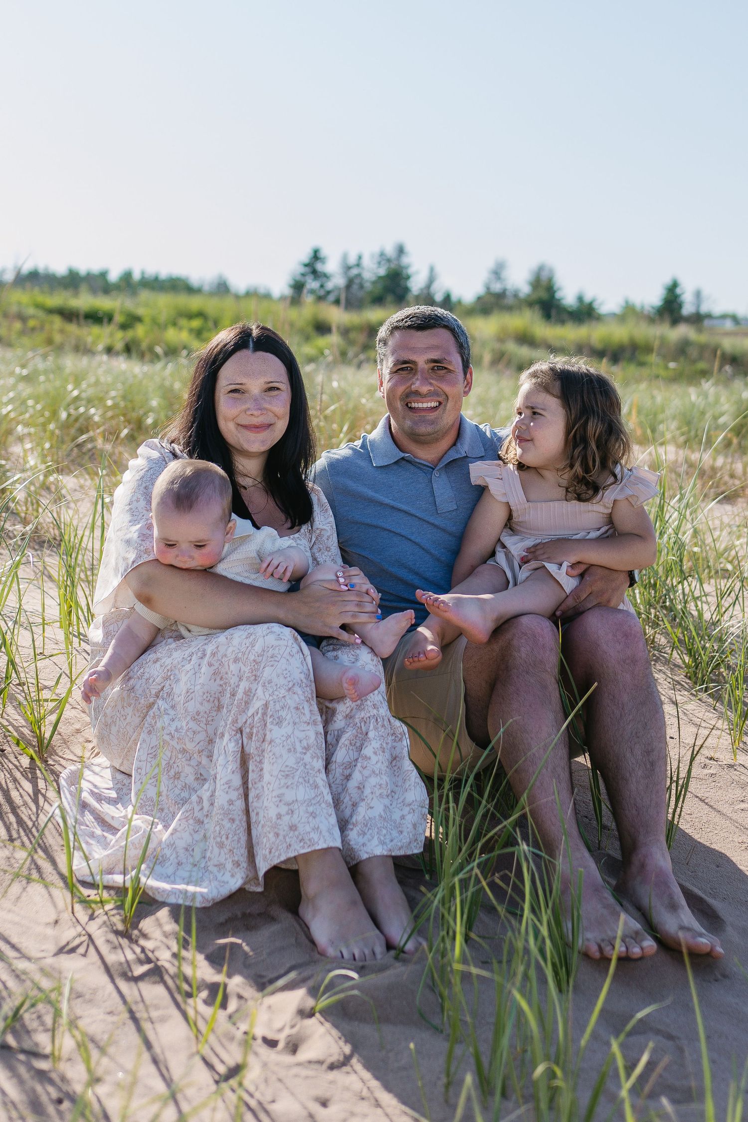 Smiling family of four sitting together on sandy beach grass dunes on a bright sunny day.