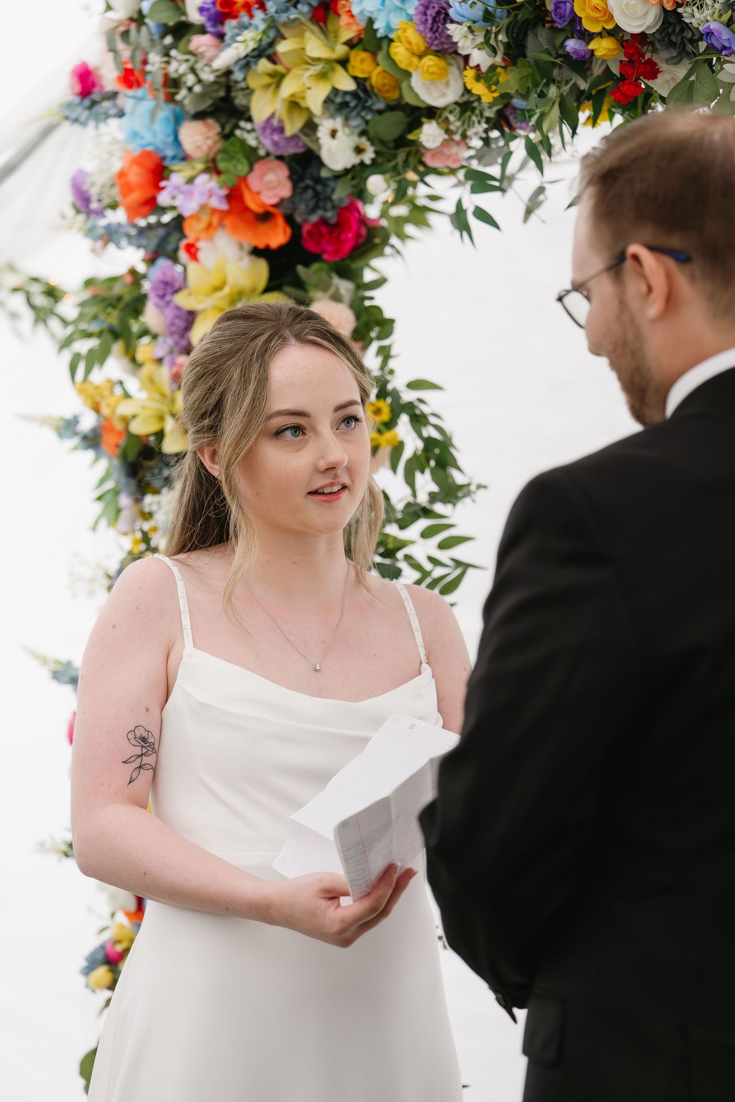 Bride in white dress reading vows under colorful floral arch at outdoor wedding ceremony.