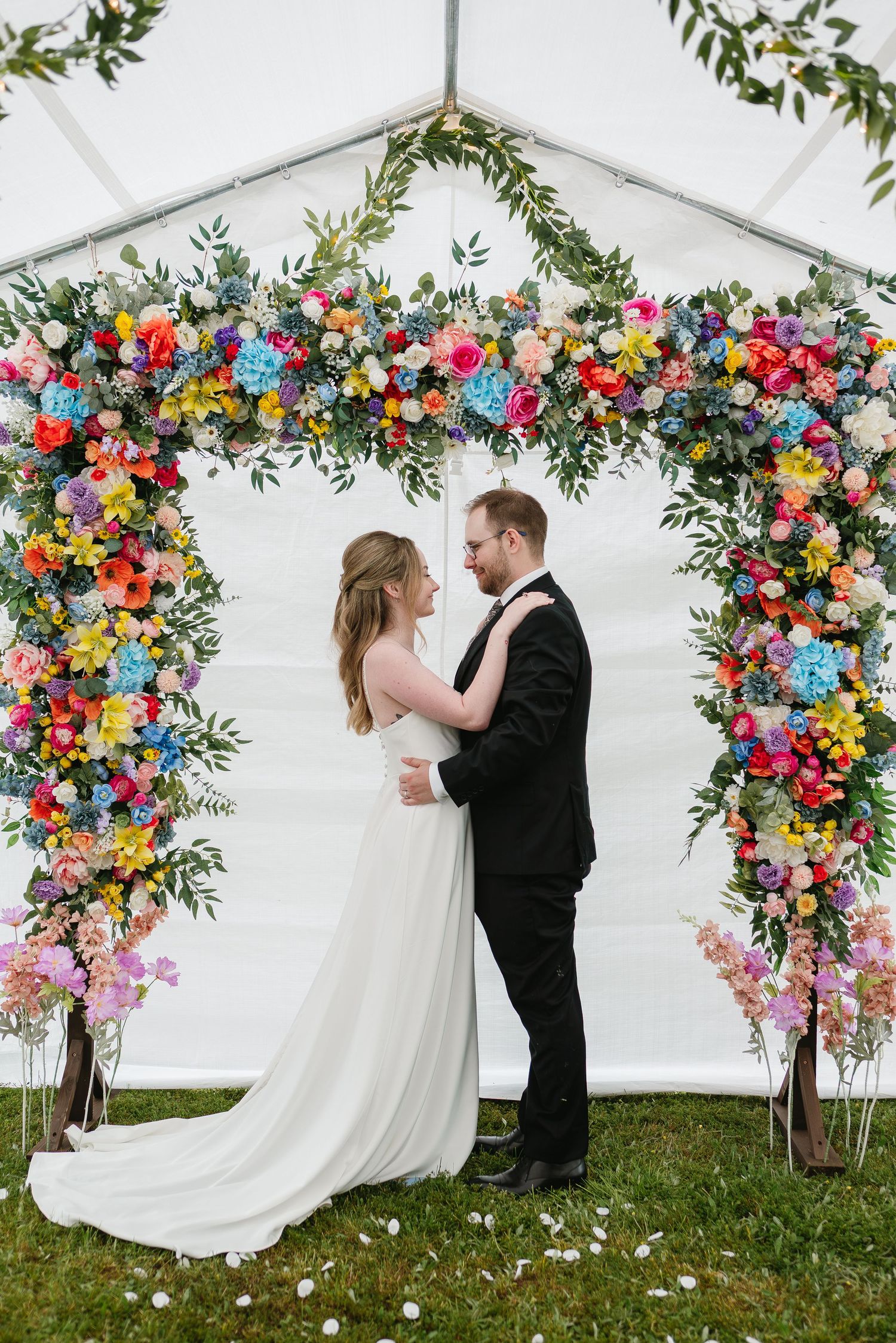 Bride and groom embracing before stunning colorful flower arch at outdoor marquee wedding.