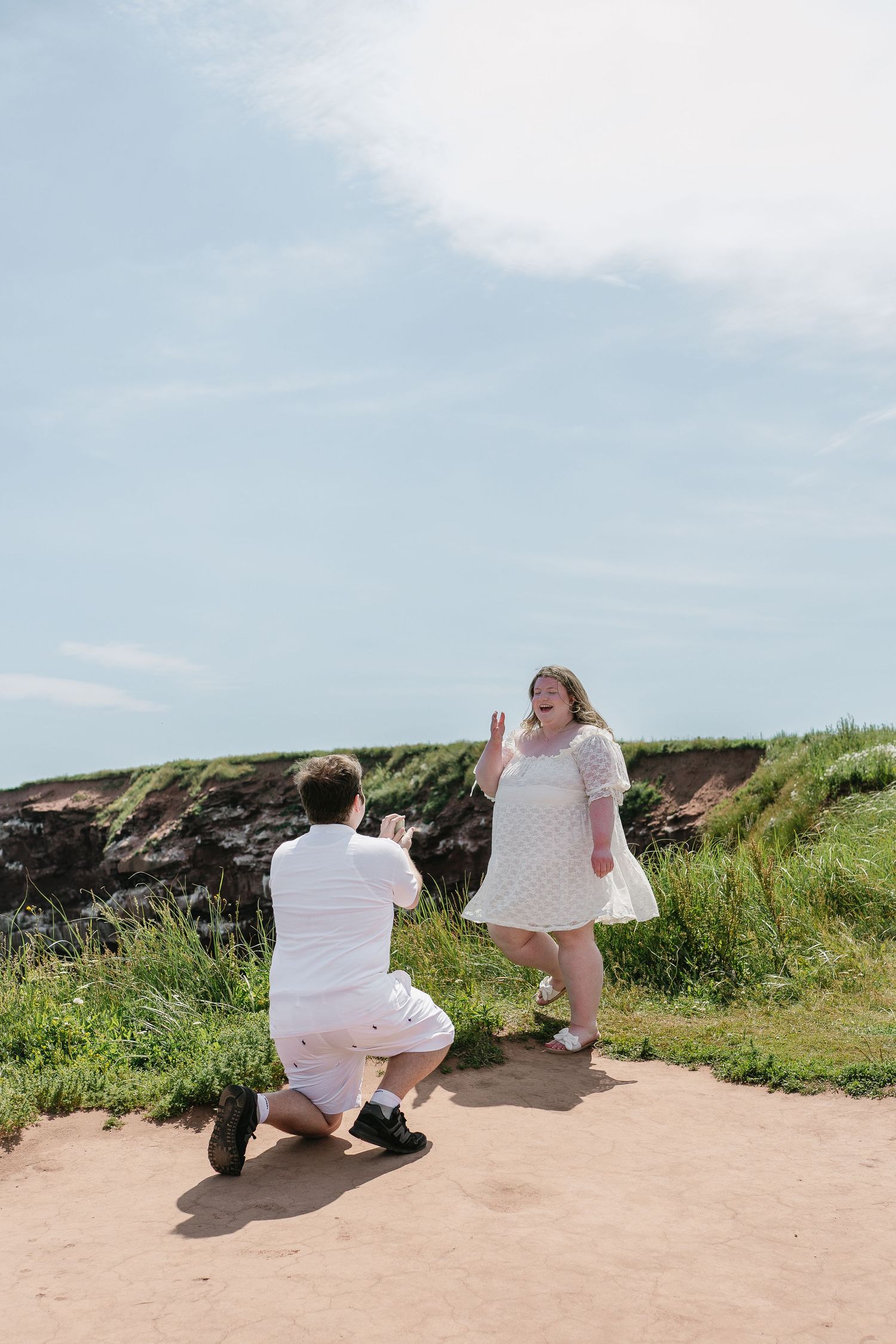 Man on one knee proposing to surprised woman in white dress at scenic coastal location.