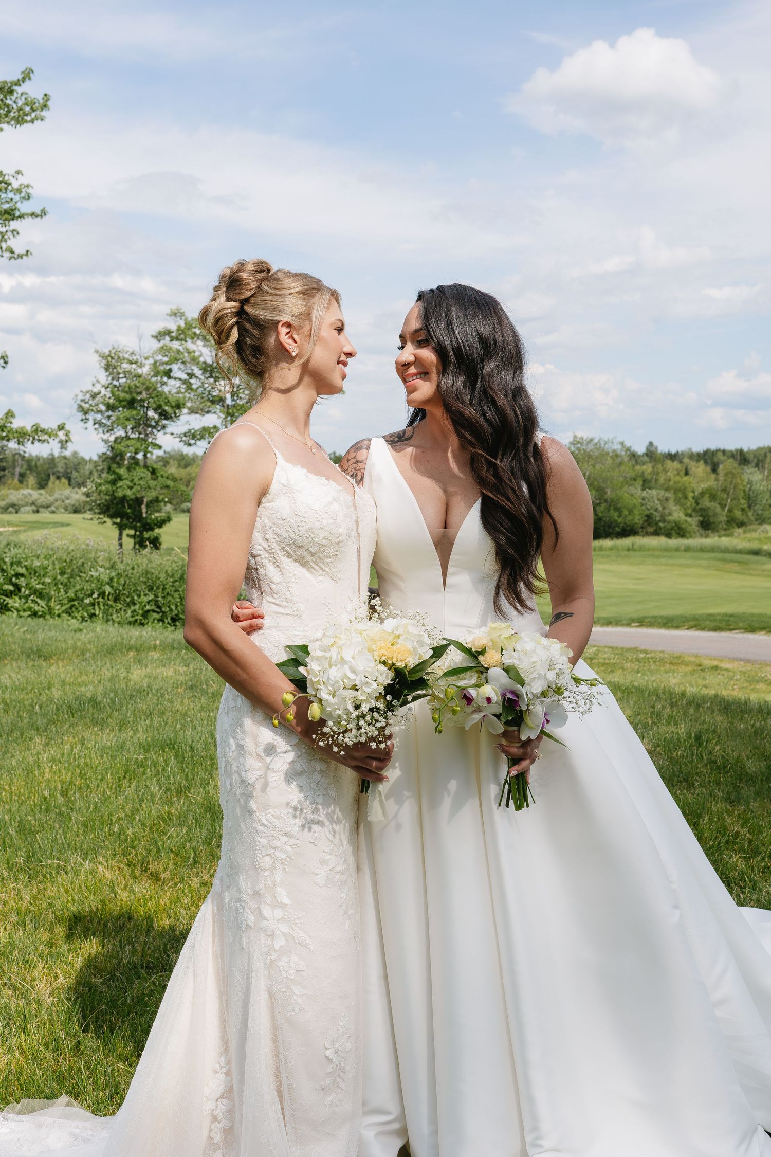 Two brides in white wedding gowns holding bouquets smile at each other in sunny outdoor setting.