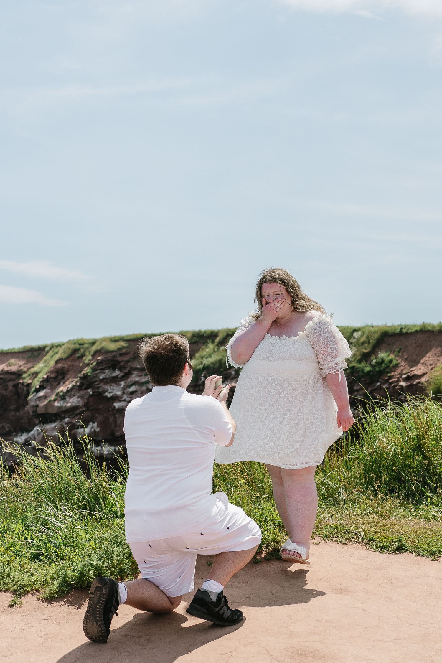 Romantic outdoor proposal with man on bended knee presenting ring to emotional woman in white dress.