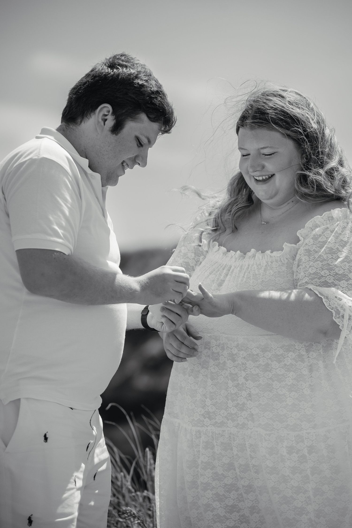 Black and white photo of a joyful couple looking down as a man places an engagement ring on her finger.