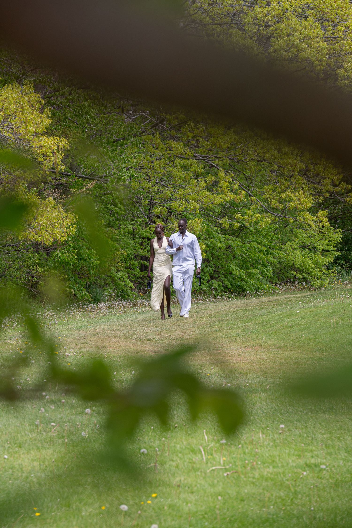 Couple walking hand-in-hand across a lush green lawn surrounded by trees in spring foliage.