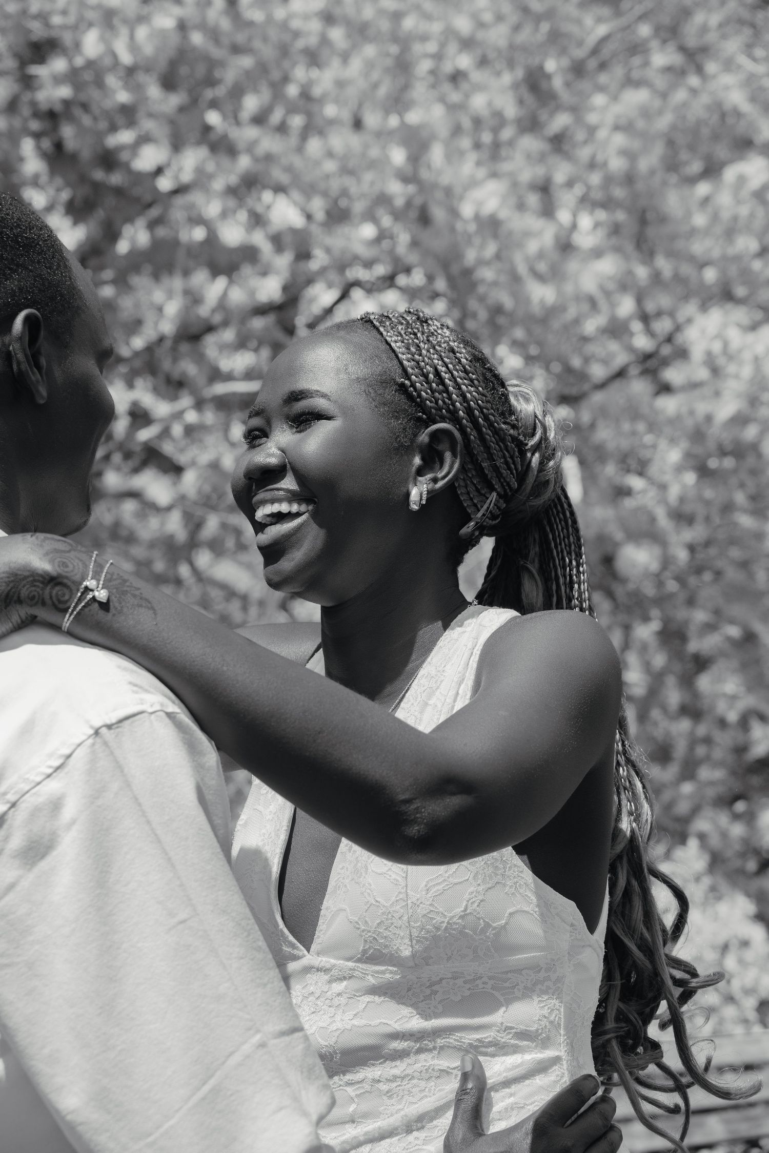Black and white photo of a laughing woman in white dress embracing her partner outdoors among trees.