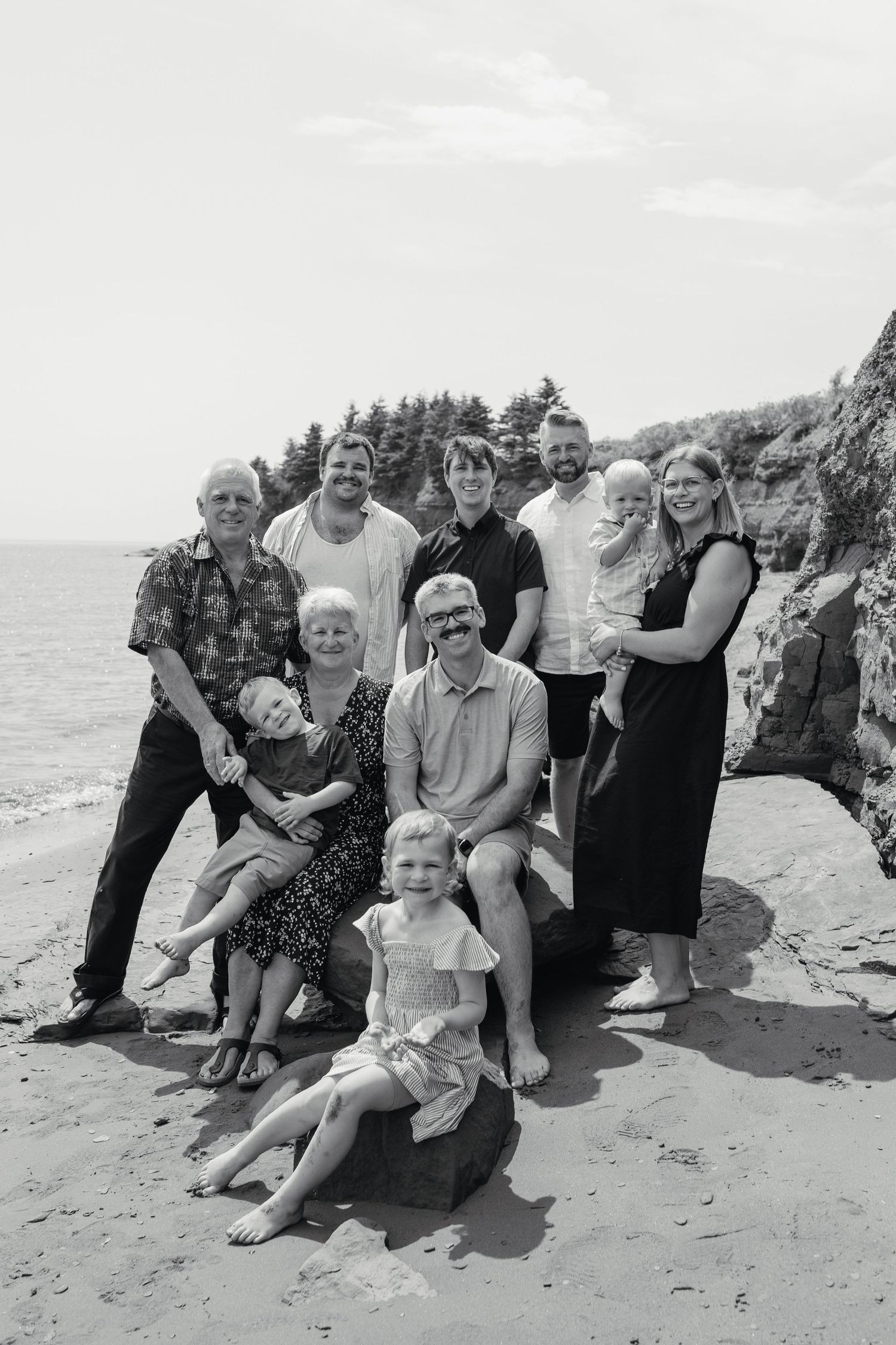 Black and white multi-generational family portrait on a sandy beach with rocky cliffs and ocean in the background.