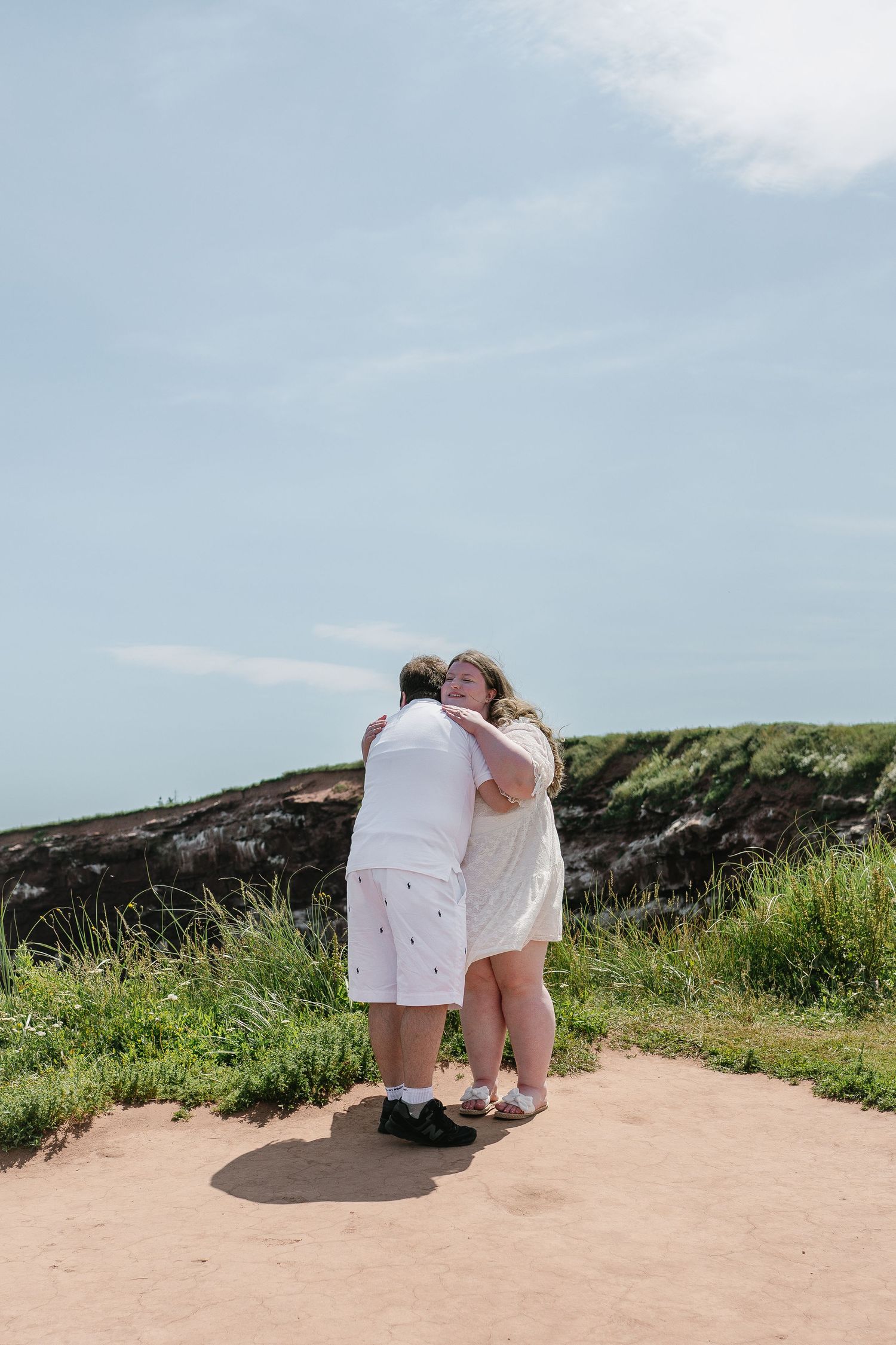 Newly engaged couple sharing a warm embrace on a sunny coastal pathway after proposal.