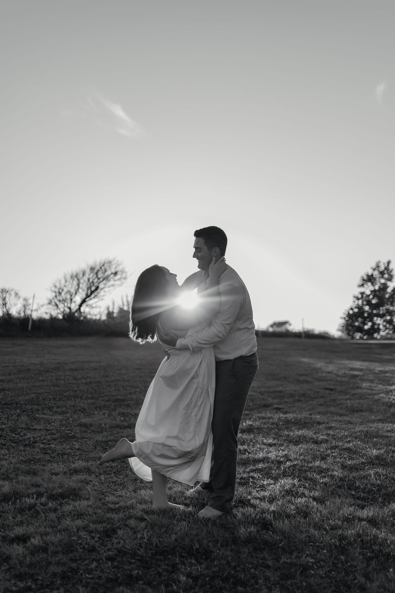 Black and white photo of a couple embracing in a field at sunset with a lens flare between them.
