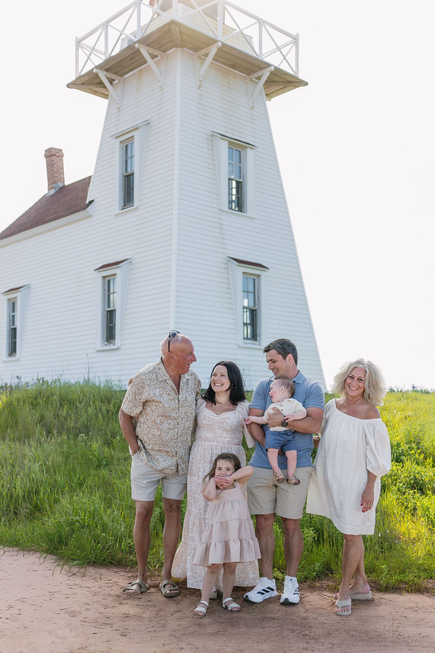 Extended family of six smiles together in front of a white lighthouse surrounded by lush green grass on a bright day.