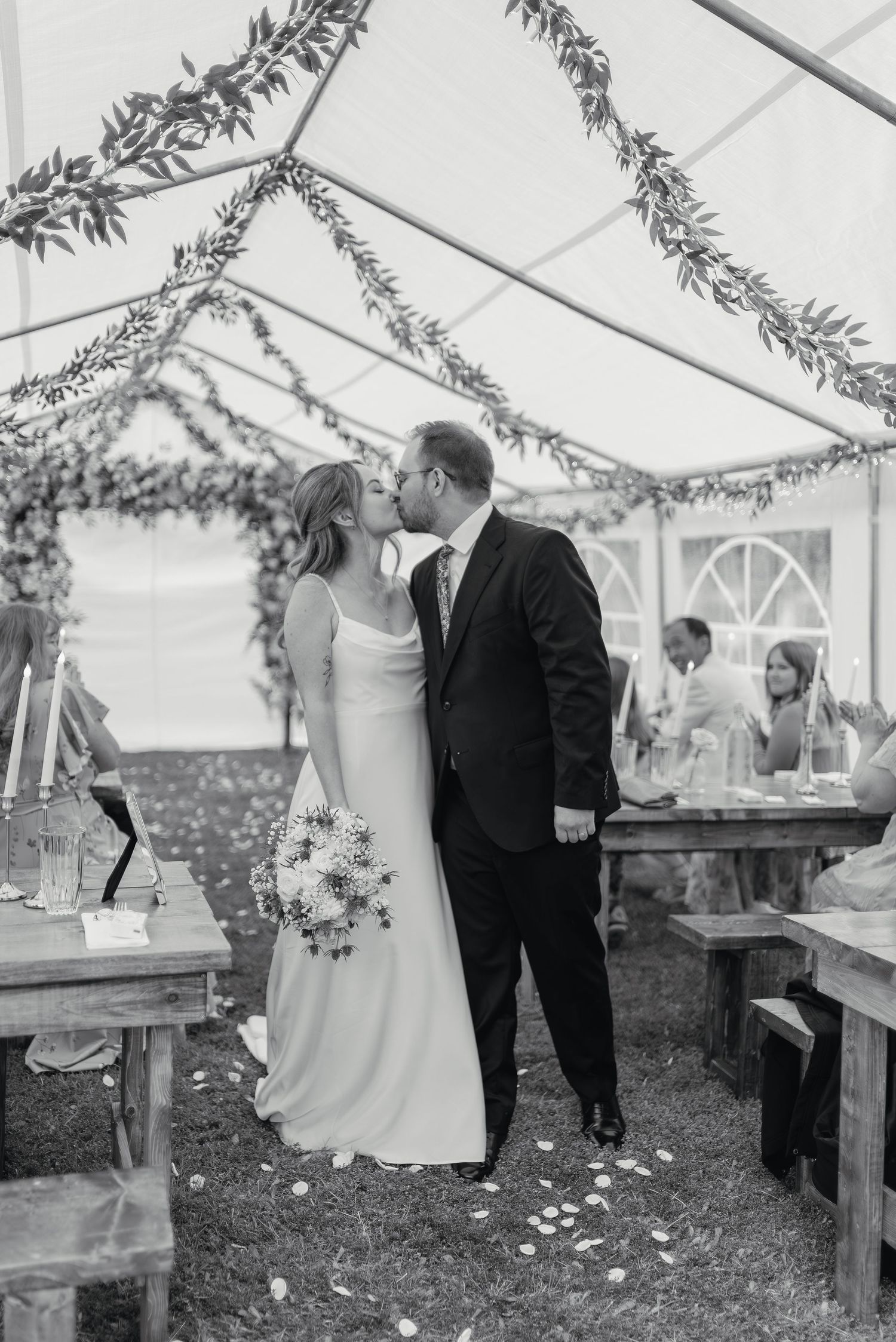 Bride and groom share first kiss under floral garland tent at outdoor wedding reception.