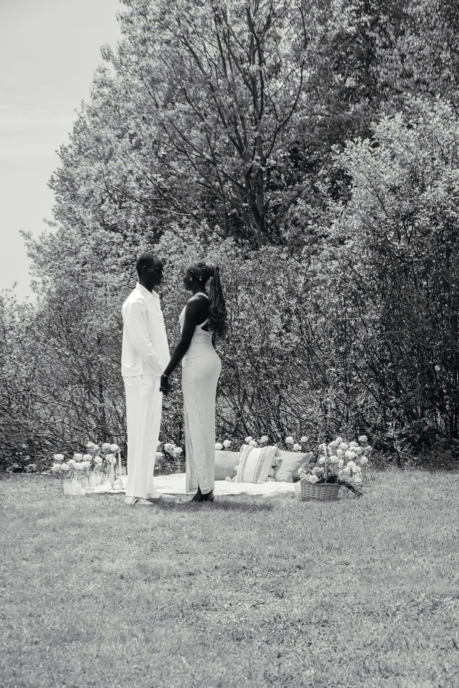 Black and white photo of a couple in white standing facing each other near floral decorations in a garden.