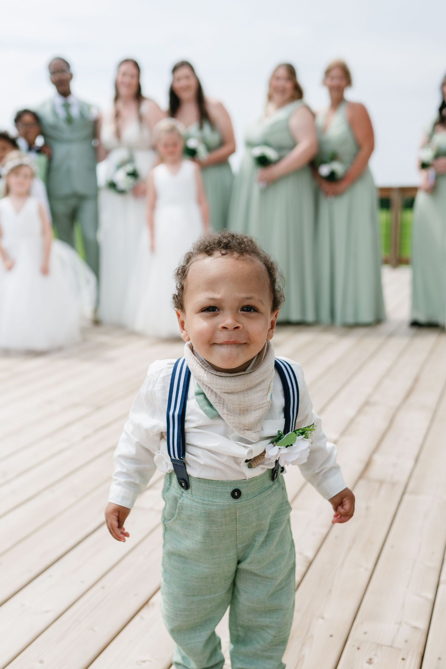 Adorable toddler ring bearer in sage pants and suspenders smiles at camera with wedding party behind him.