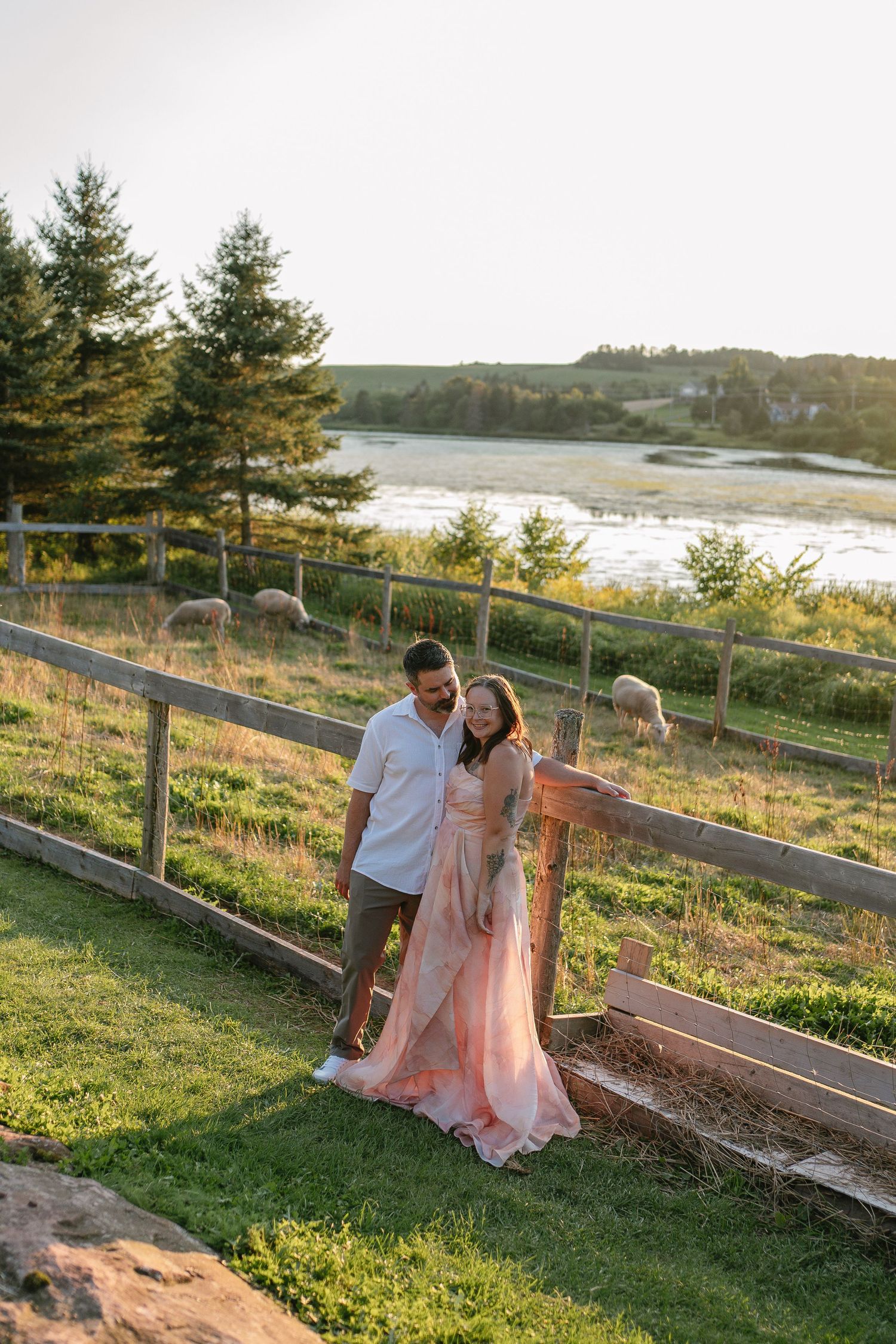 Couple posing by rustic wooden fence at golden hour, woman in pink gown, scenic river and green landscape behind them.