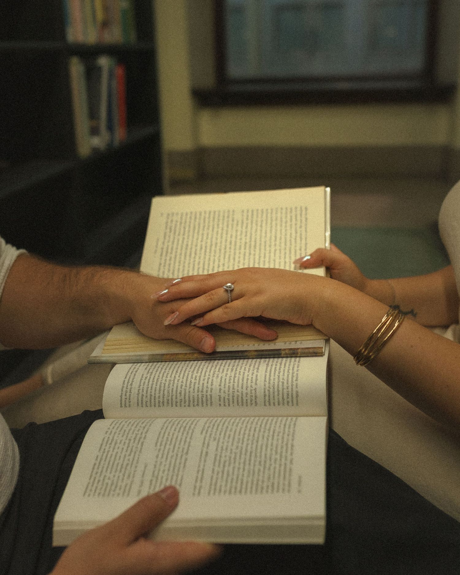 Two hands rest together on an open book with gold rings visible on their fingers in an intimate reading moment.