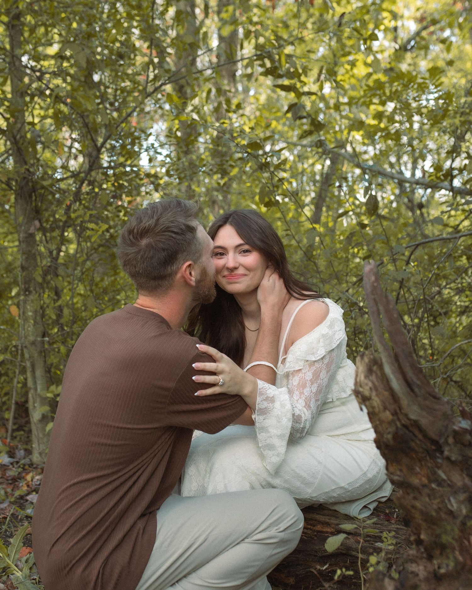 A couple shares a tender moment in a sunlit forest setting with green foliage in the background.