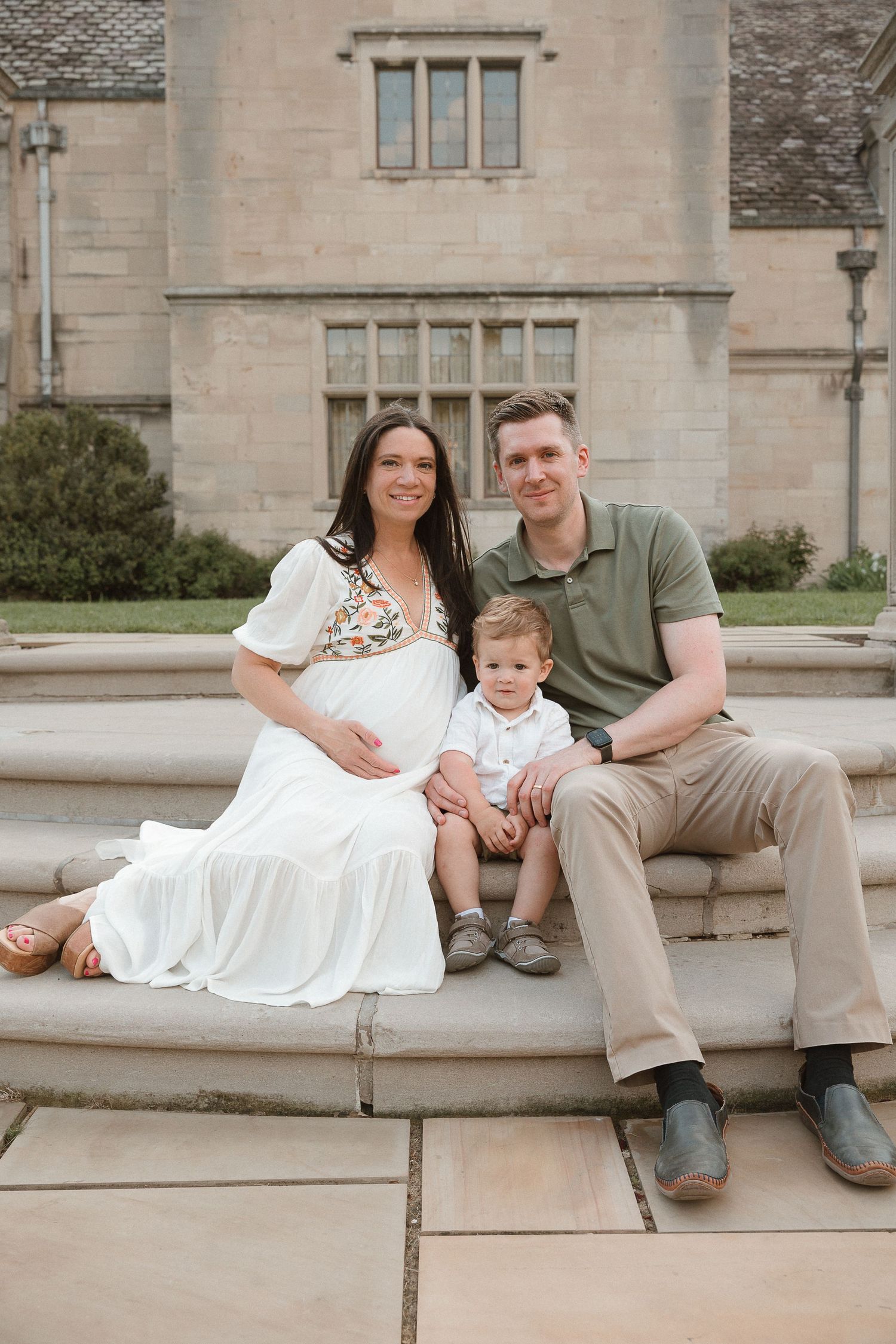 Cinematic maternity portrait with soft film edit, family of three sitting by a fountain in natural tones and peaceful light.