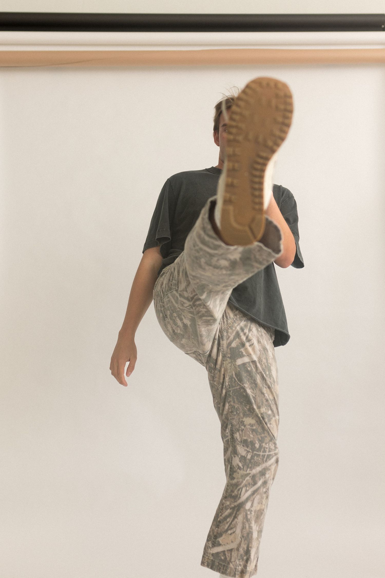 Person in black t-shirt and patterned pants kicks leg up high against white background.