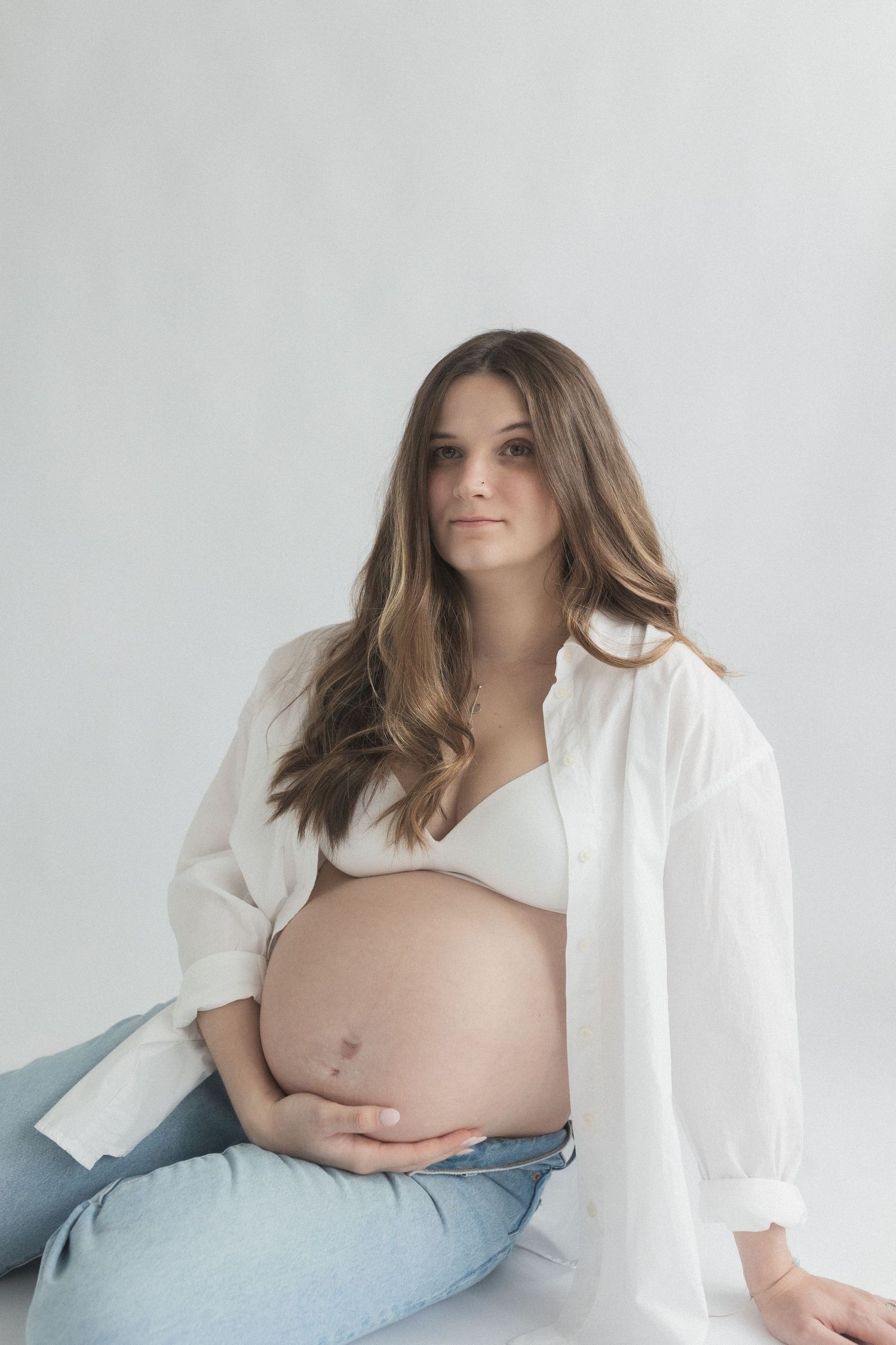 Side view maternity photo with woman in white button-down shirt against minimalist background.
