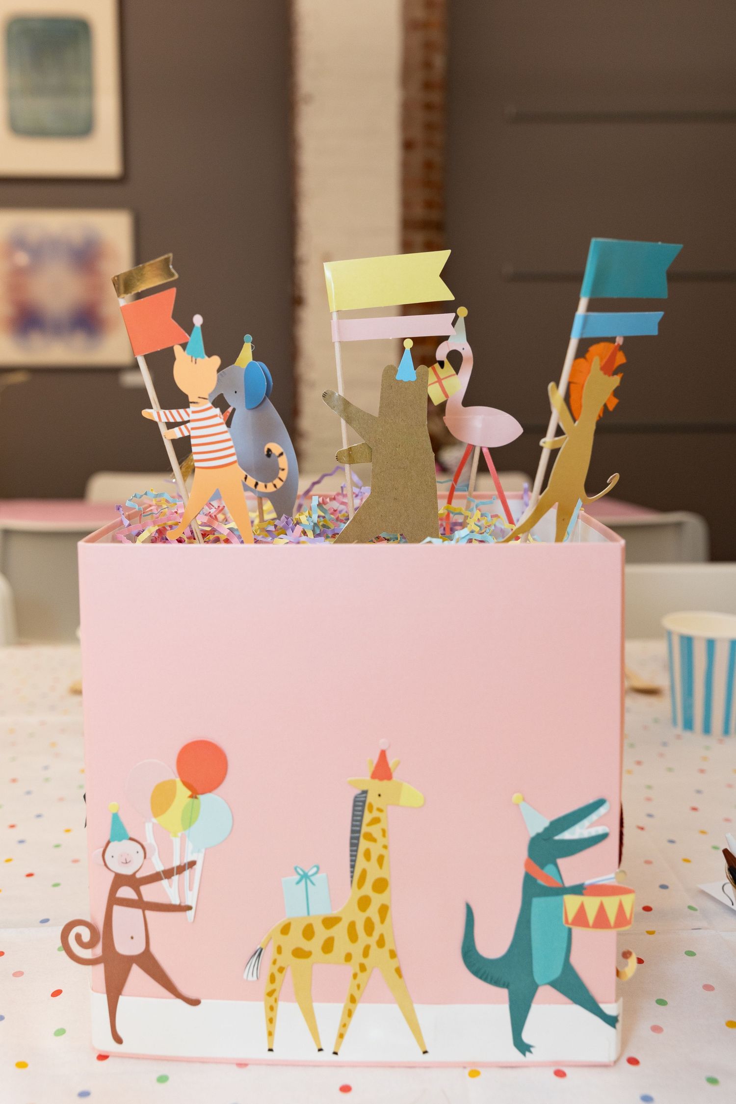 Whimsical pink birthday cake decorated with colorful paper animals and flag toppers featuring a monkey, giraffe and rabbit.