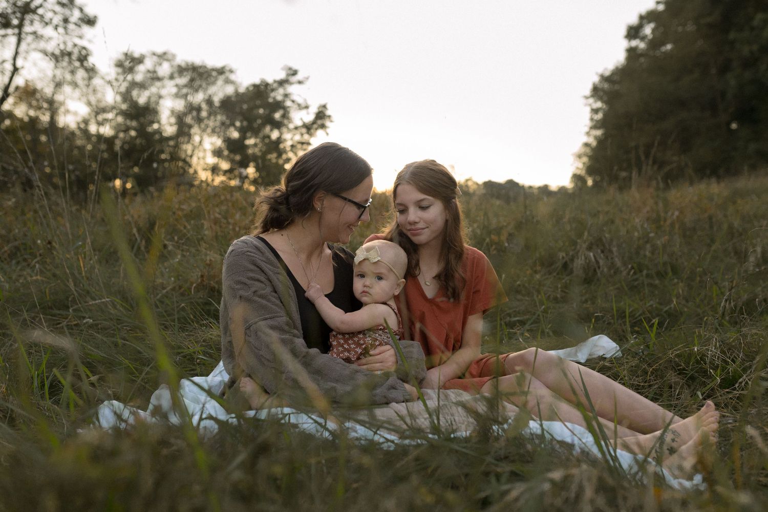 Three people share a tender moment while sitting on a blanket in a field at sunset.