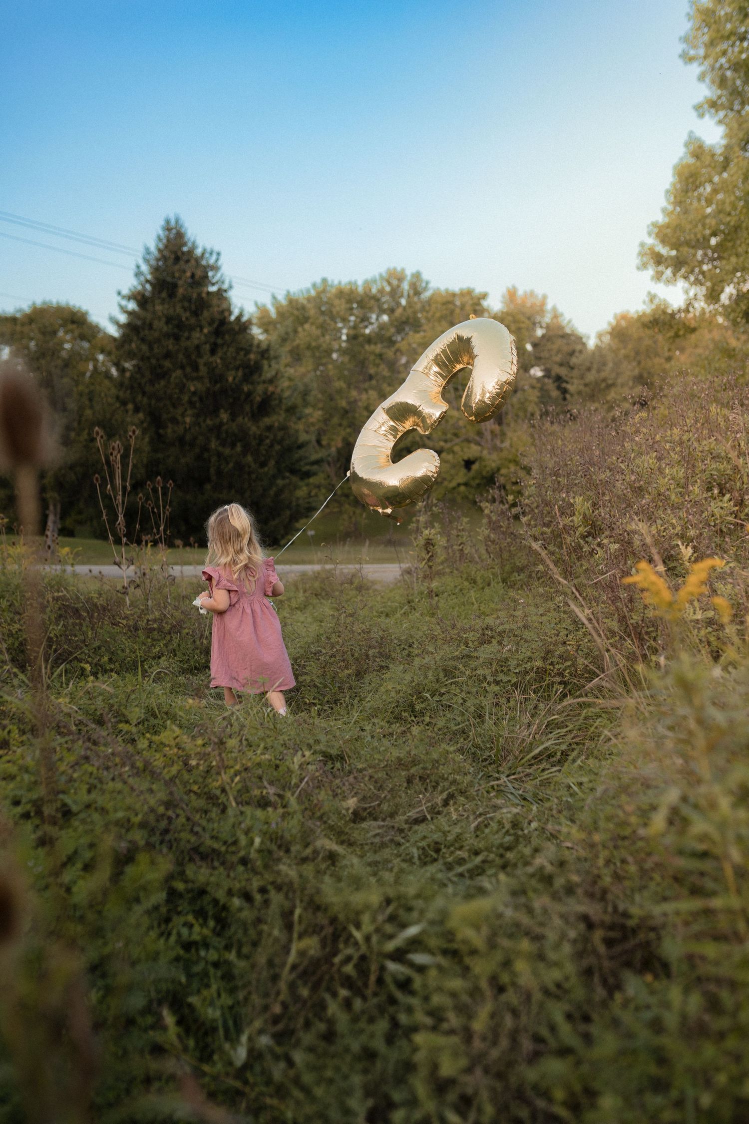 Child in pink dress watches large number balloon float in rural field.
