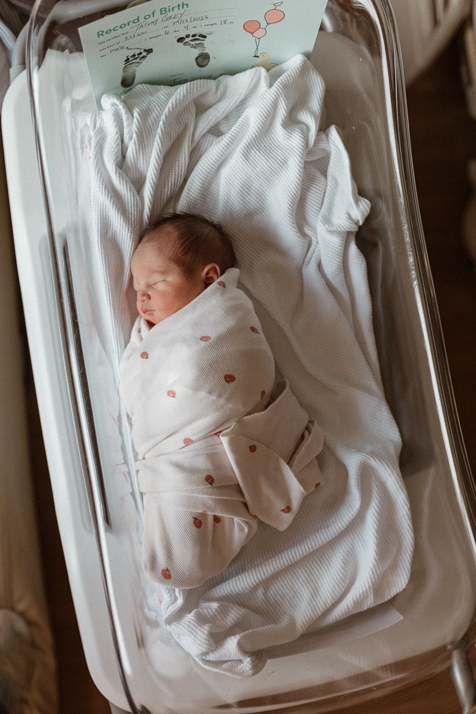 Newborn baby sleeps in hospital bassinet wrapped in white polka dot swaddle next to birth record.