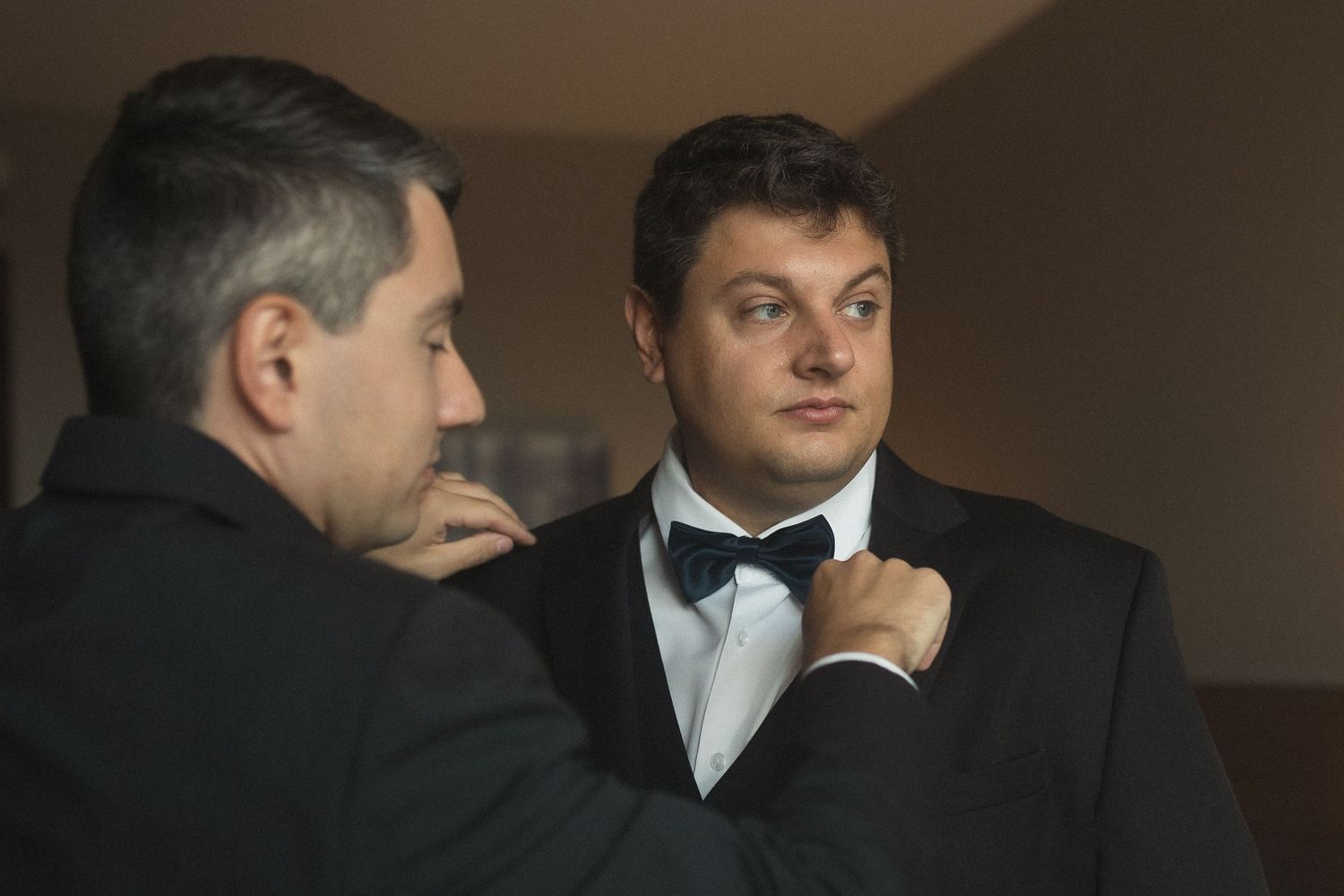 Two men in formal attire adjusting a bowtie before a wedding ceremony.
