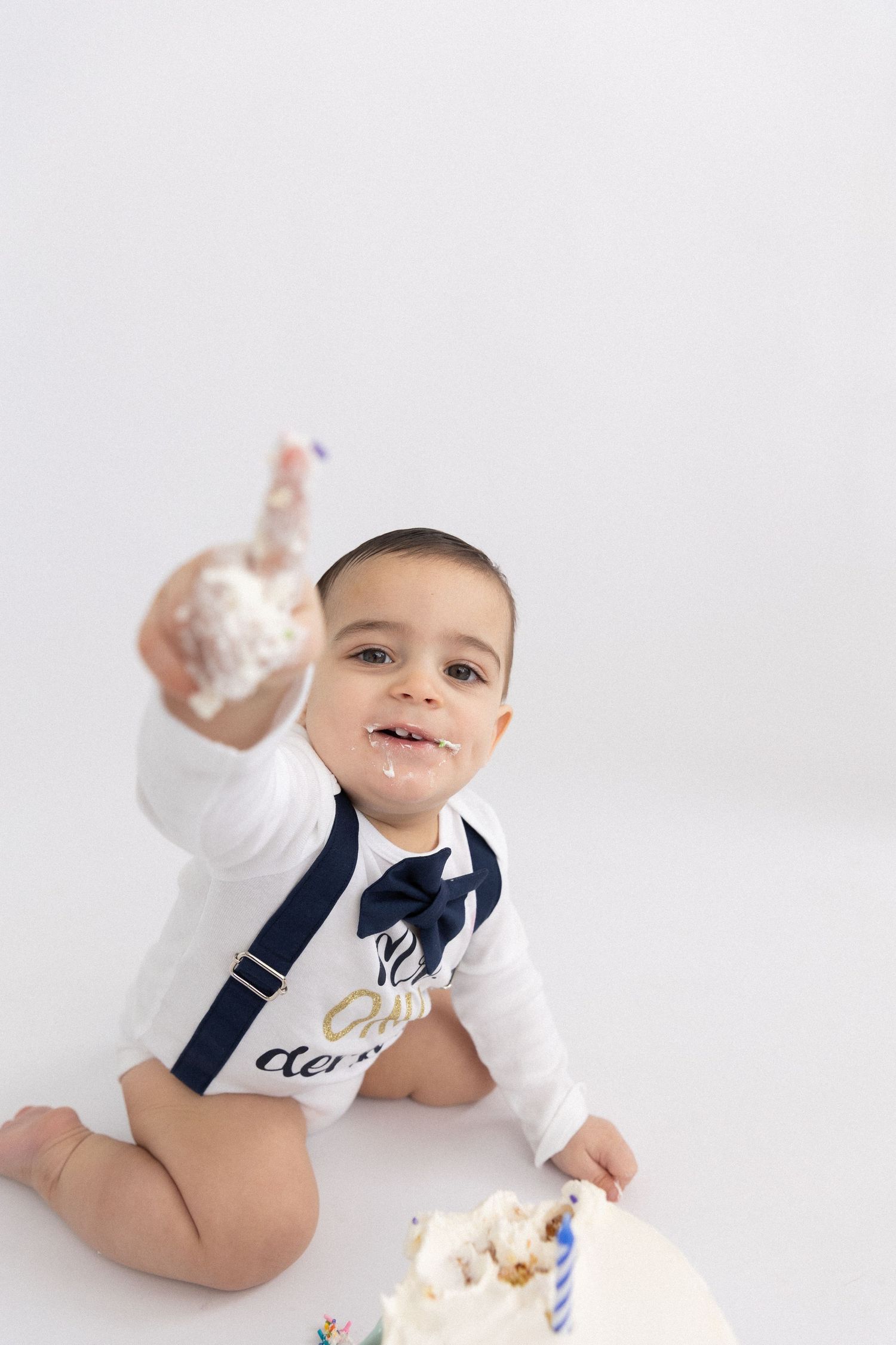 First birthday cake smash session with a smiling baby boy in a bowtie, bright studio setup, milestone photography for toddlers.