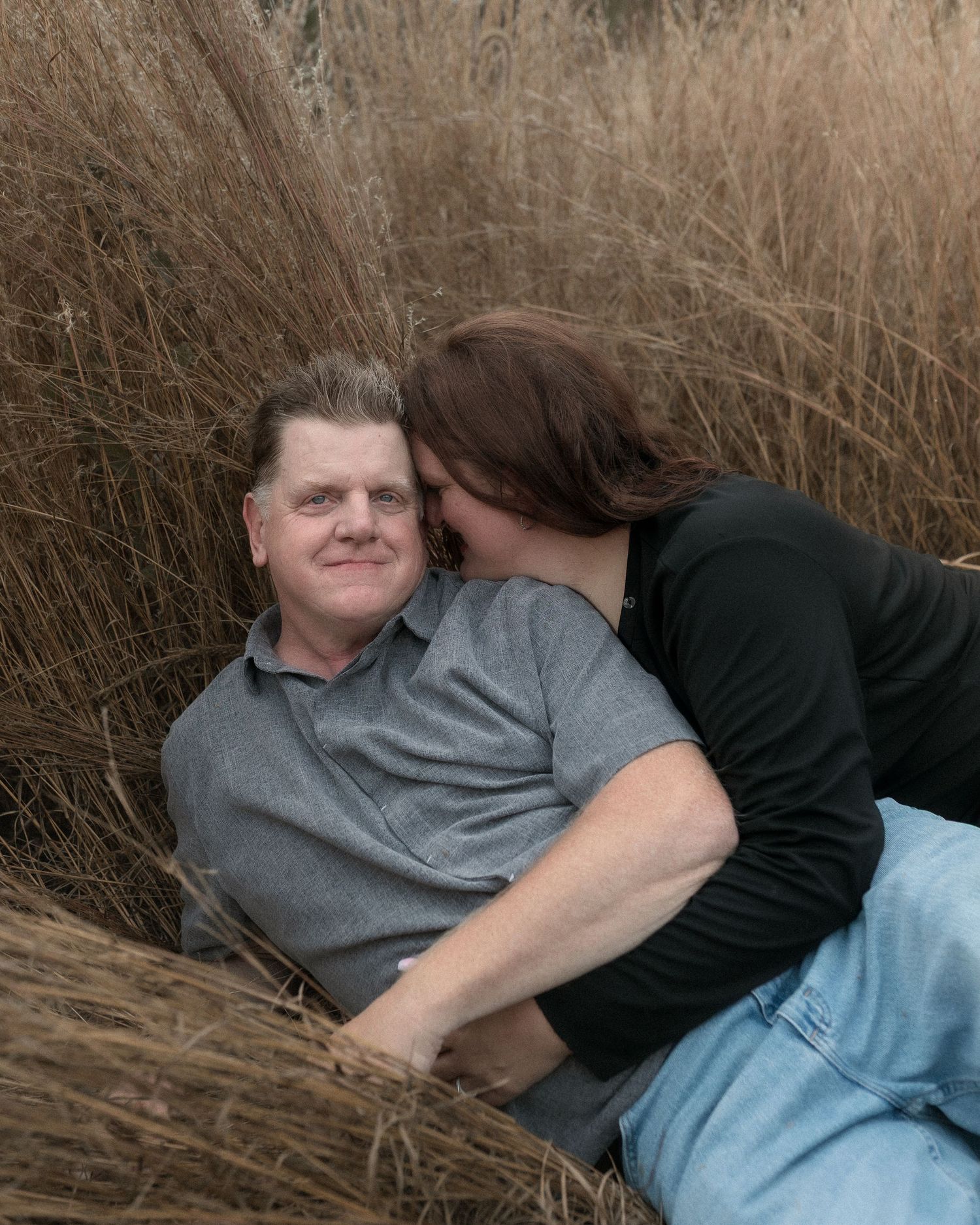 A person lies in tall brown grass wearing a gray shirt and dark jacket on a cloudy day.
