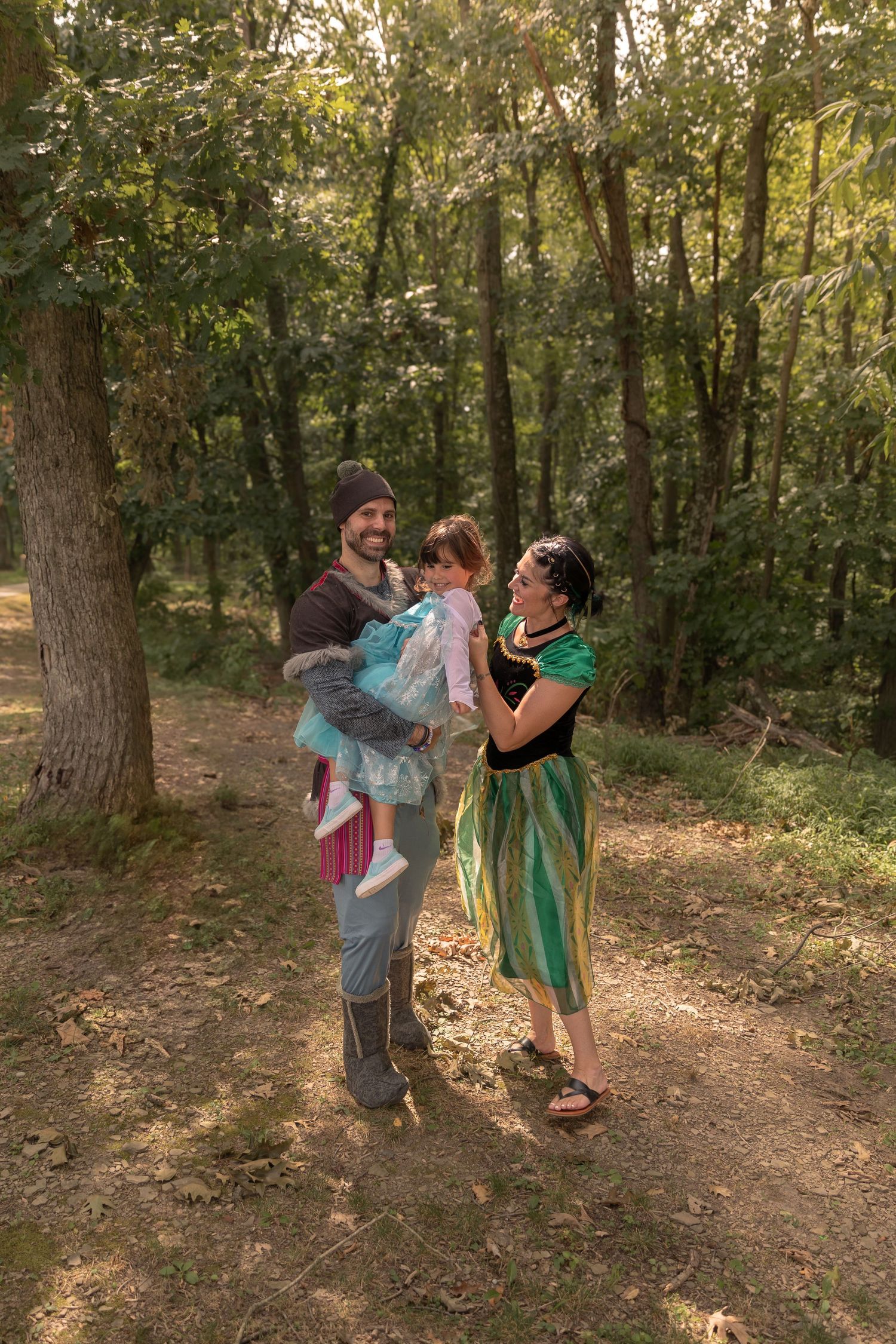 Three people in fairy tale costumes pose together in a sunlit wooded area.