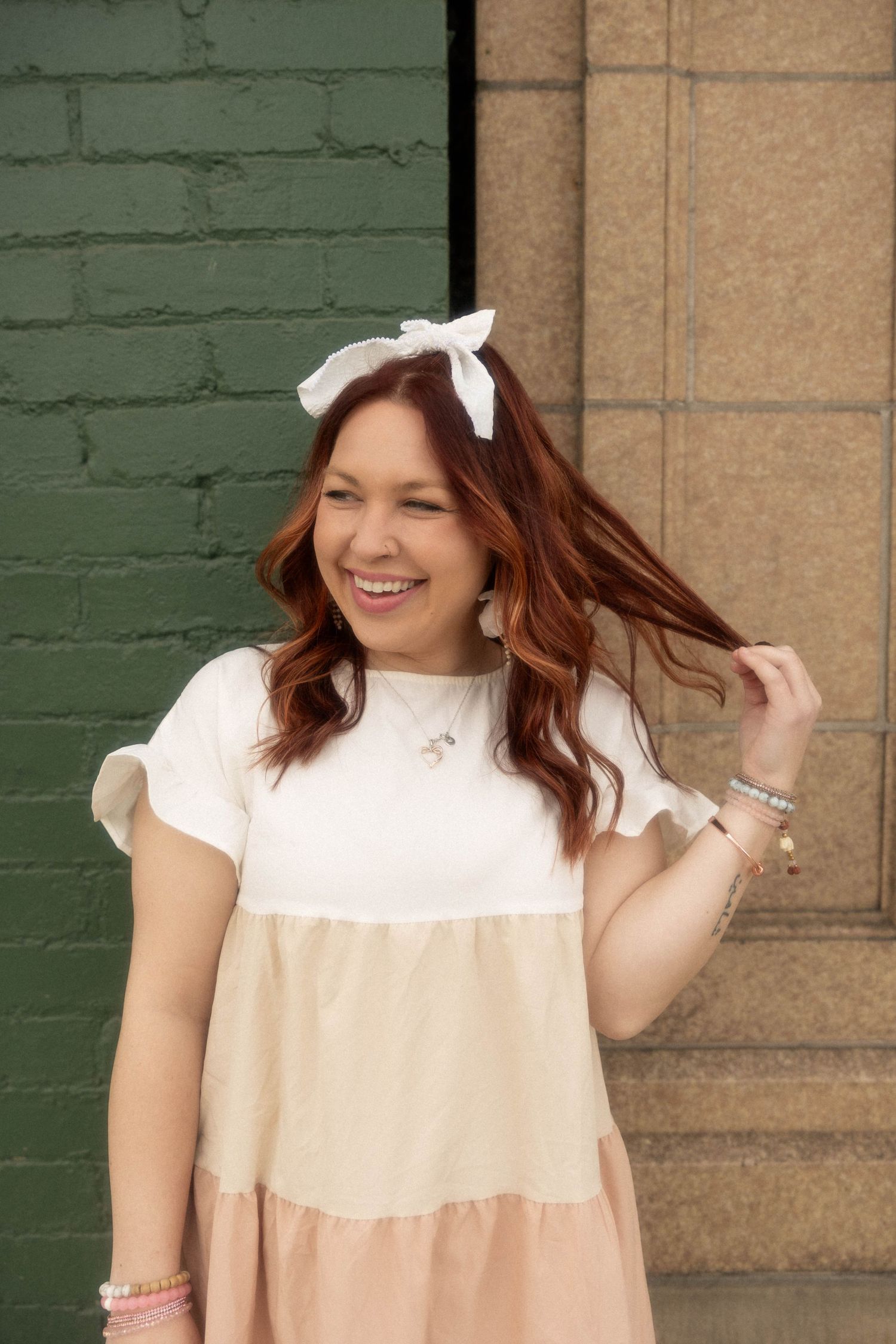 Joyful pose in peach tiered dress with white bow accessory against green brick wall.