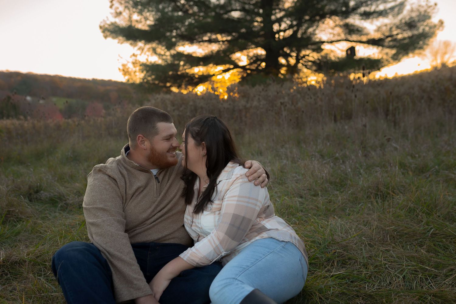 Couple sharing an intimate moment in a grassy field during golden hour sunset with pine trees in background.