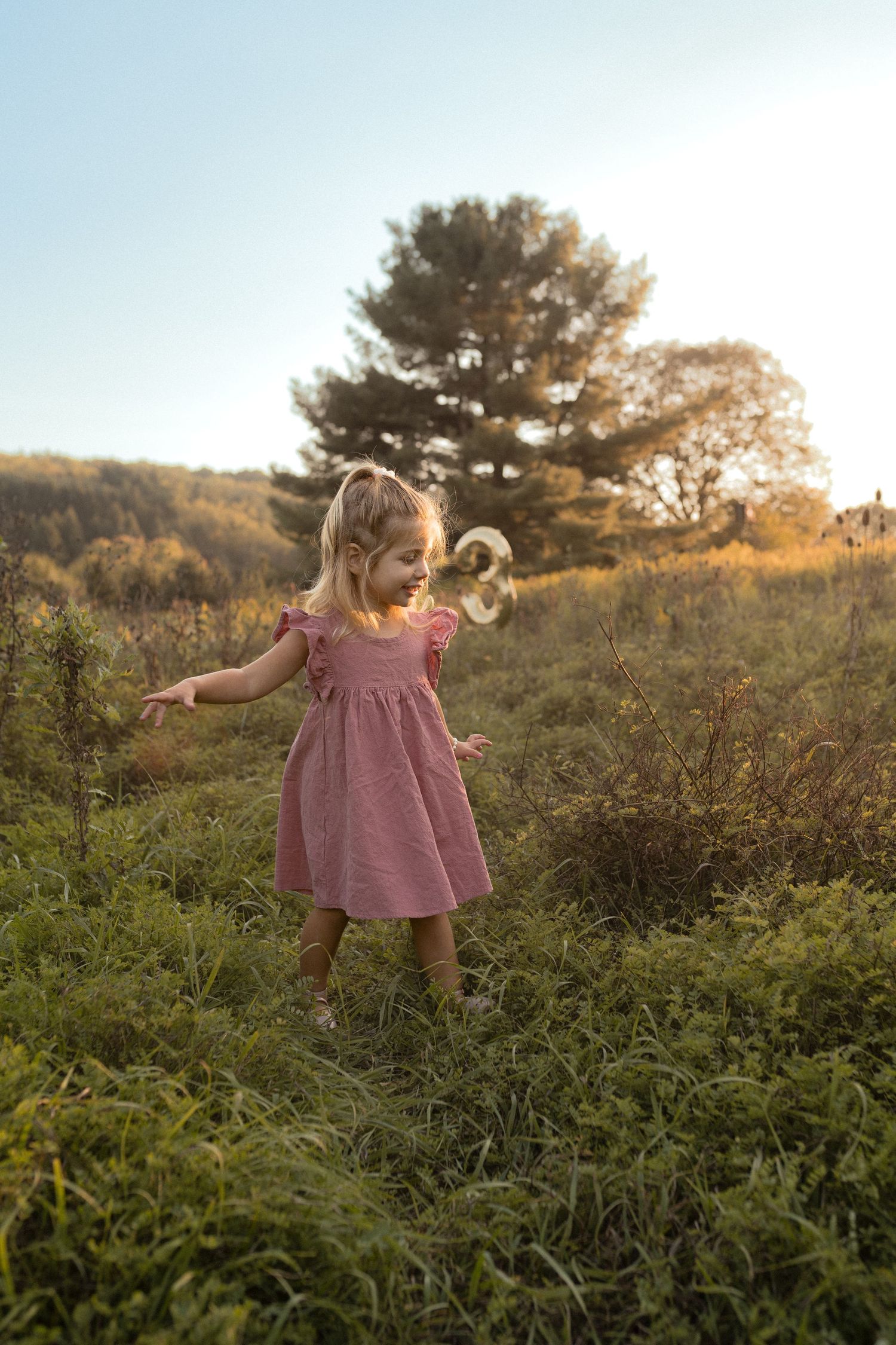 Young child in pink dress twirls playfully in meadow at sunset.