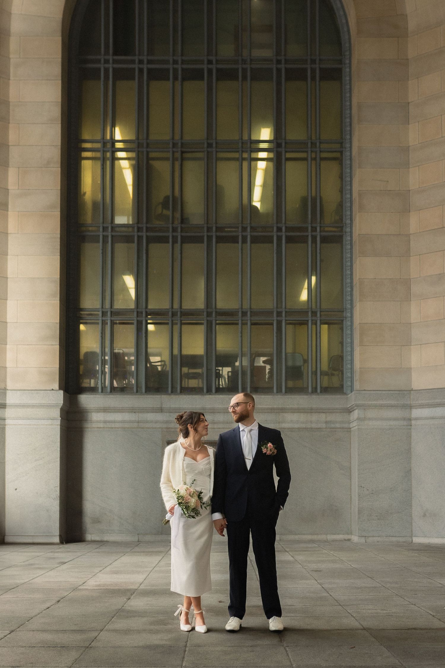 Wedding couple standing in front of large arched window at elegant venue.