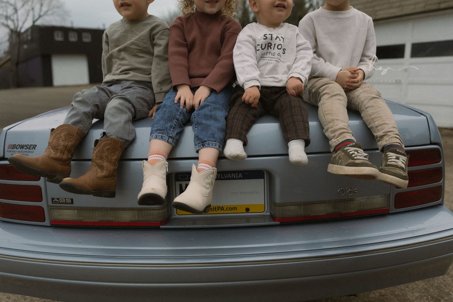 Children’s legs dangling off a vintage car bumper, cozy neutral tones in a creative lifestyle family session.