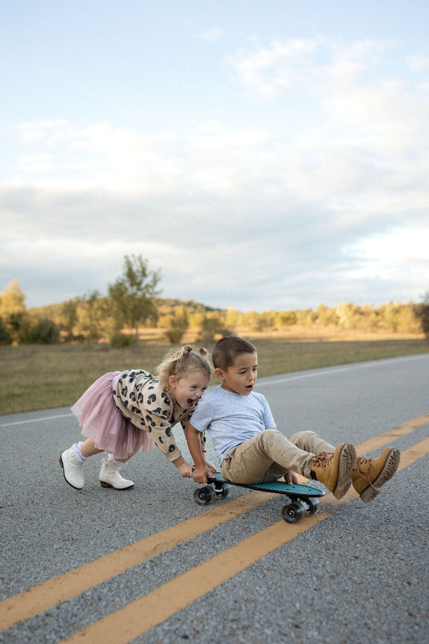 Two children playing with a skateboard on a rural road at sunset.