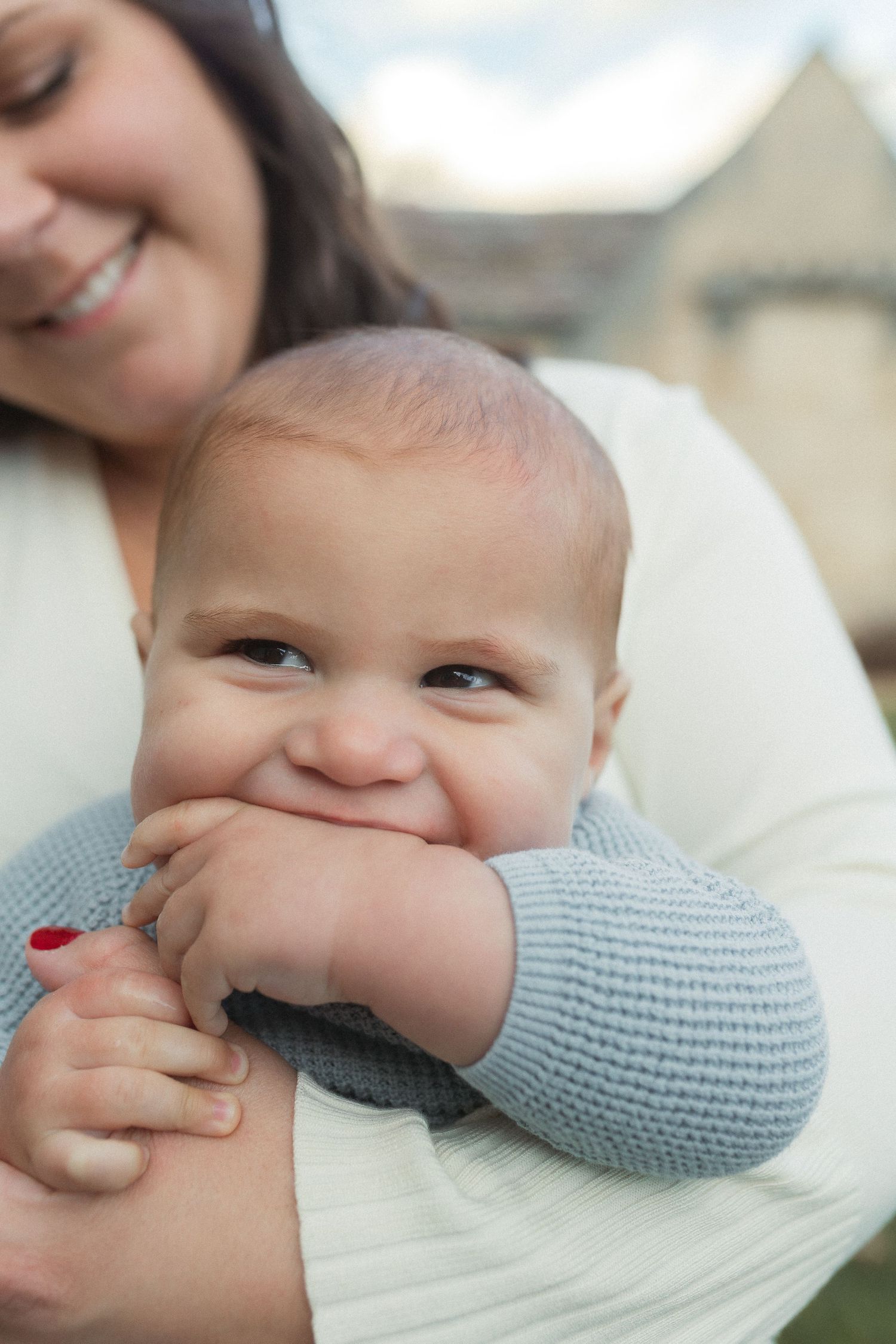 Baby with a sweet smile wearing a gray sweater being held tenderly outdoors.