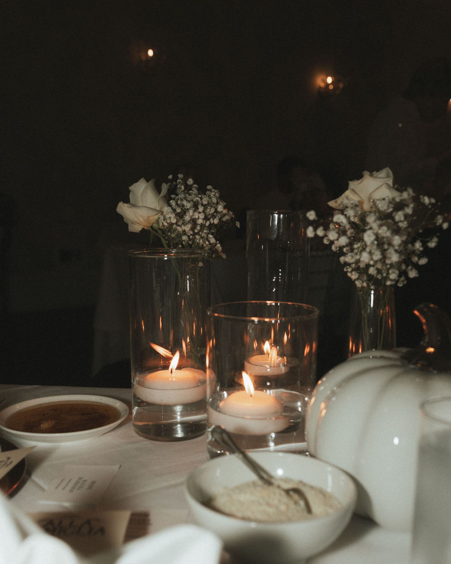 Candlelit table setting with white ceramic dishes and delicate floral arrangements in glass vases creating a romantic ambiance.