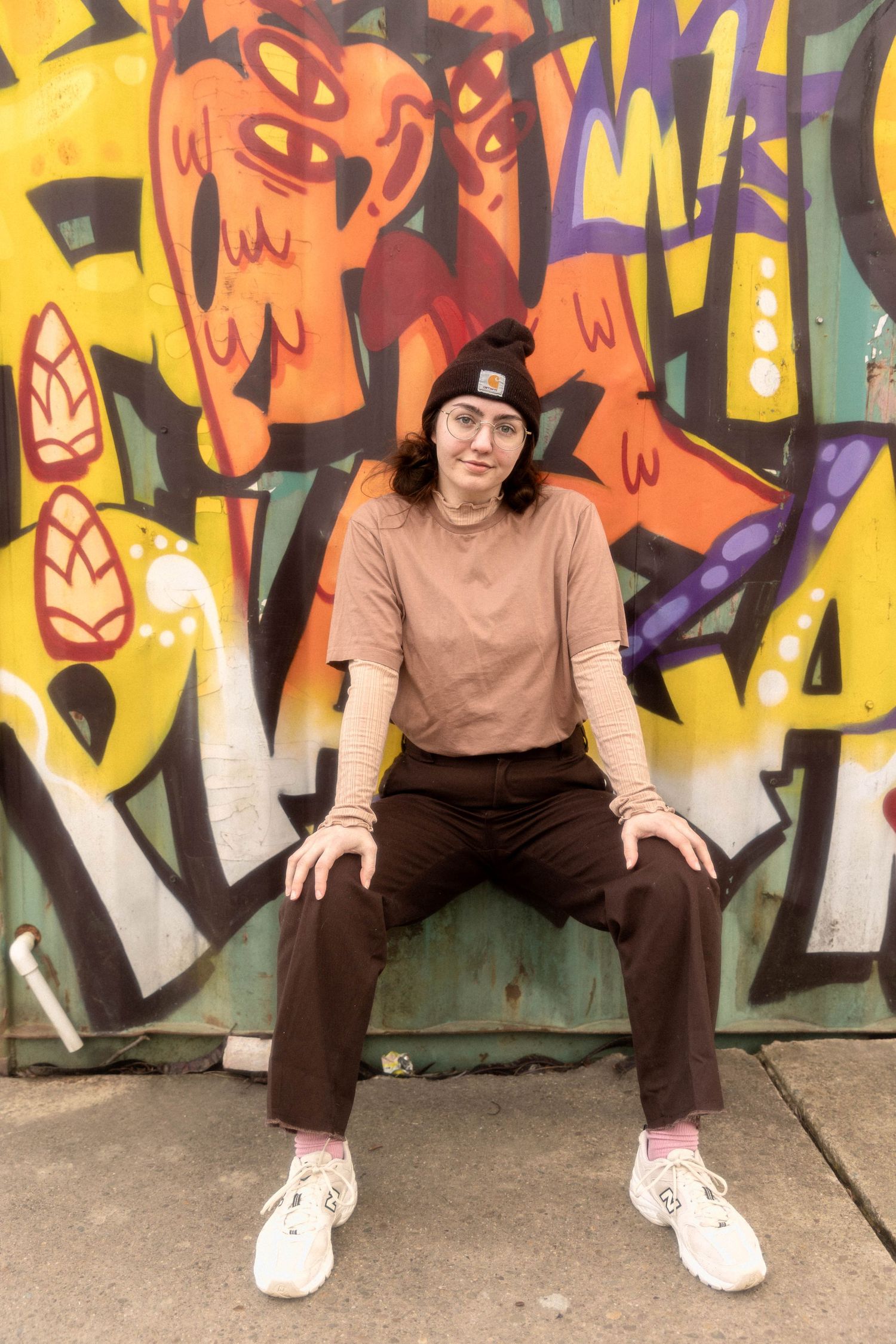 Person in beanie sits against vibrant graffiti wall wearing casual streetwear.