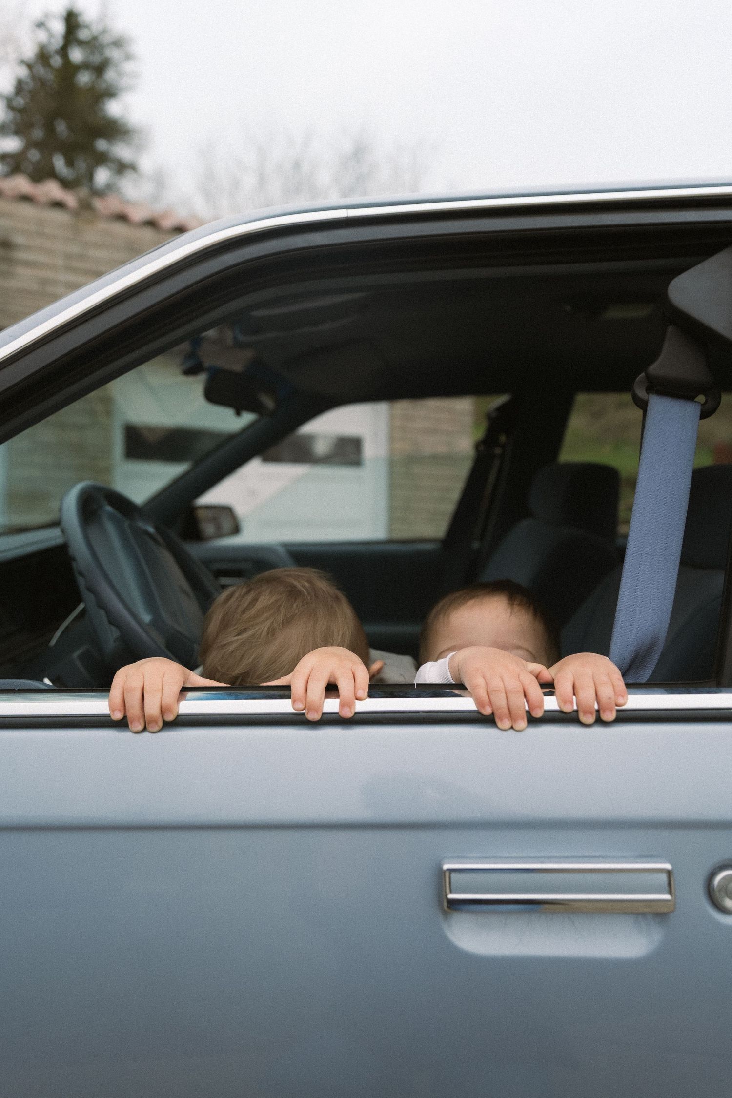Two small figures playfully peek out the window of a parked gray car.