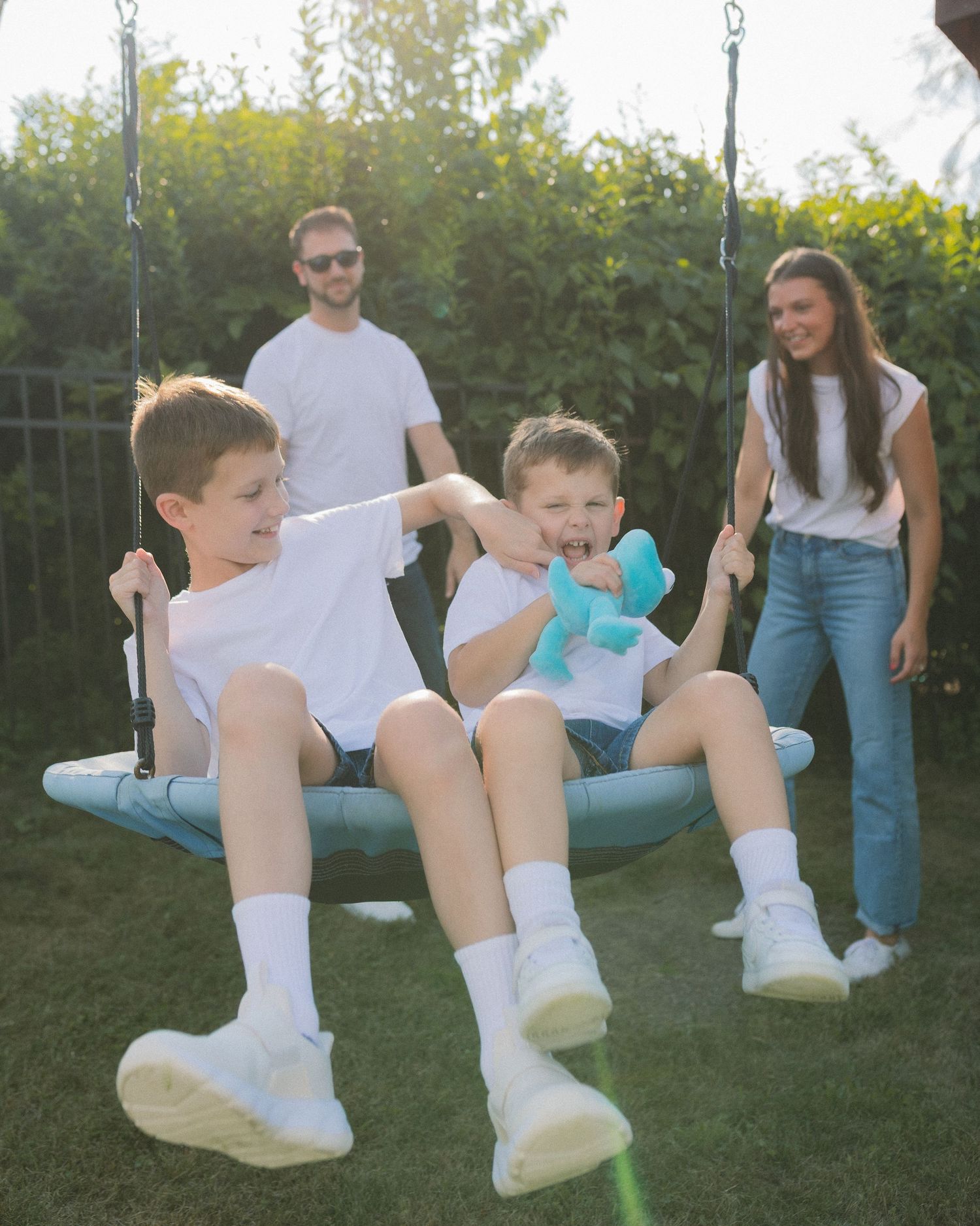 Children enjoying outdoor play on a saucer swing while adults watch from behind in a sunny backyard setting.