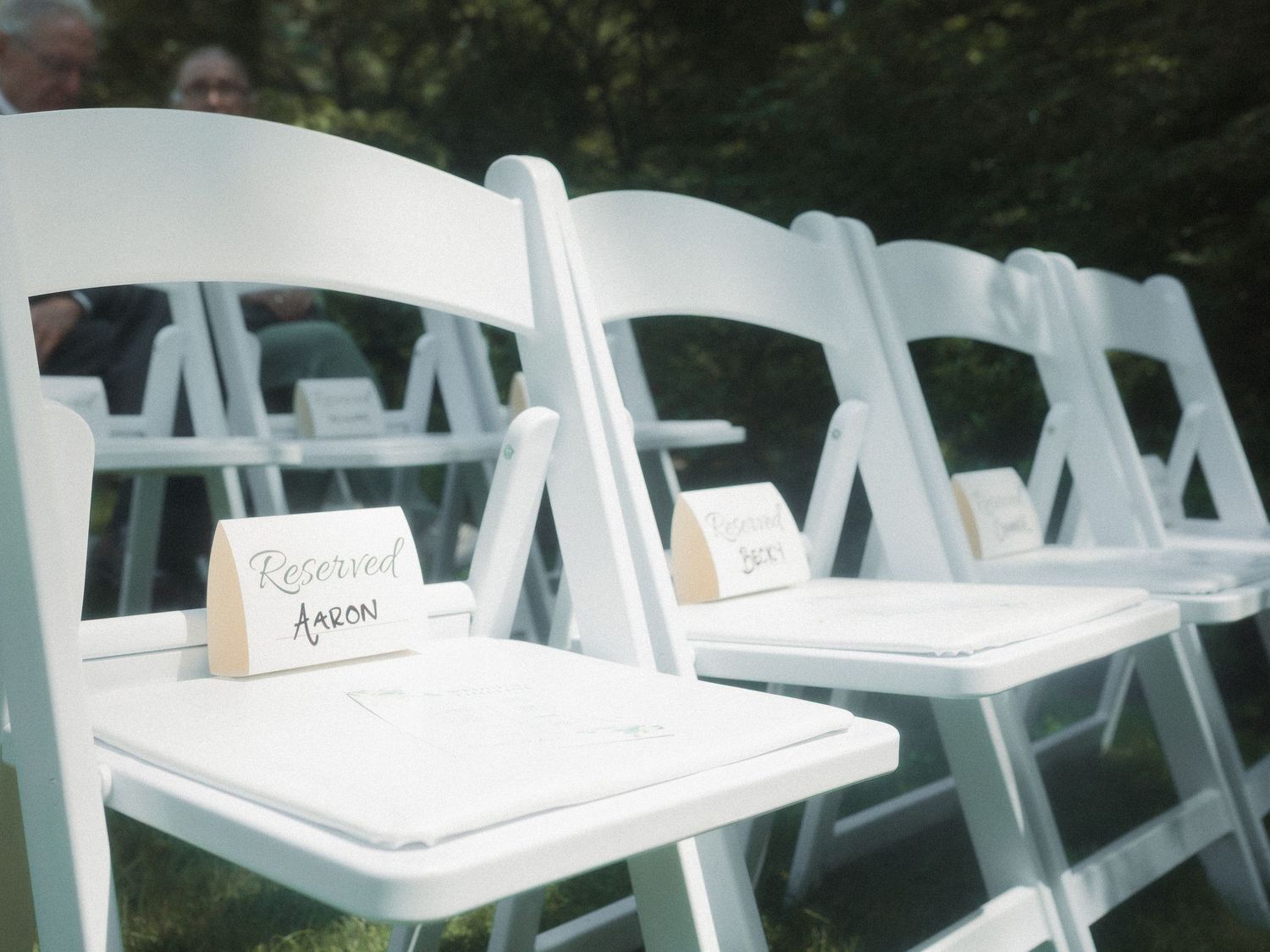 Row of white folding chairs set up outdoors with reserved signs on seats at a wedding or event.