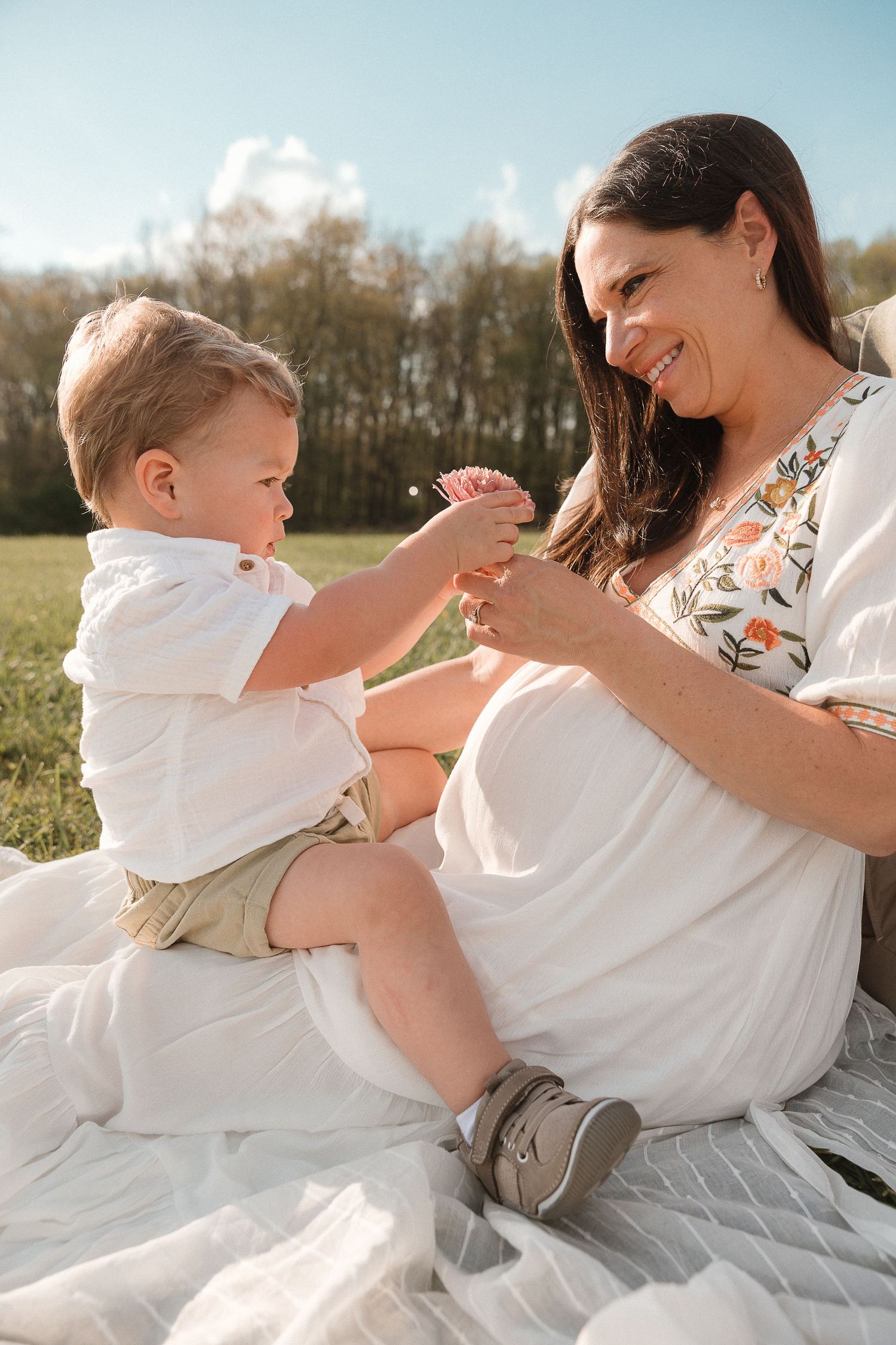 Mother and toddler sitting together on a white blanket, touching belly in joyful anticipation during maternity photos.