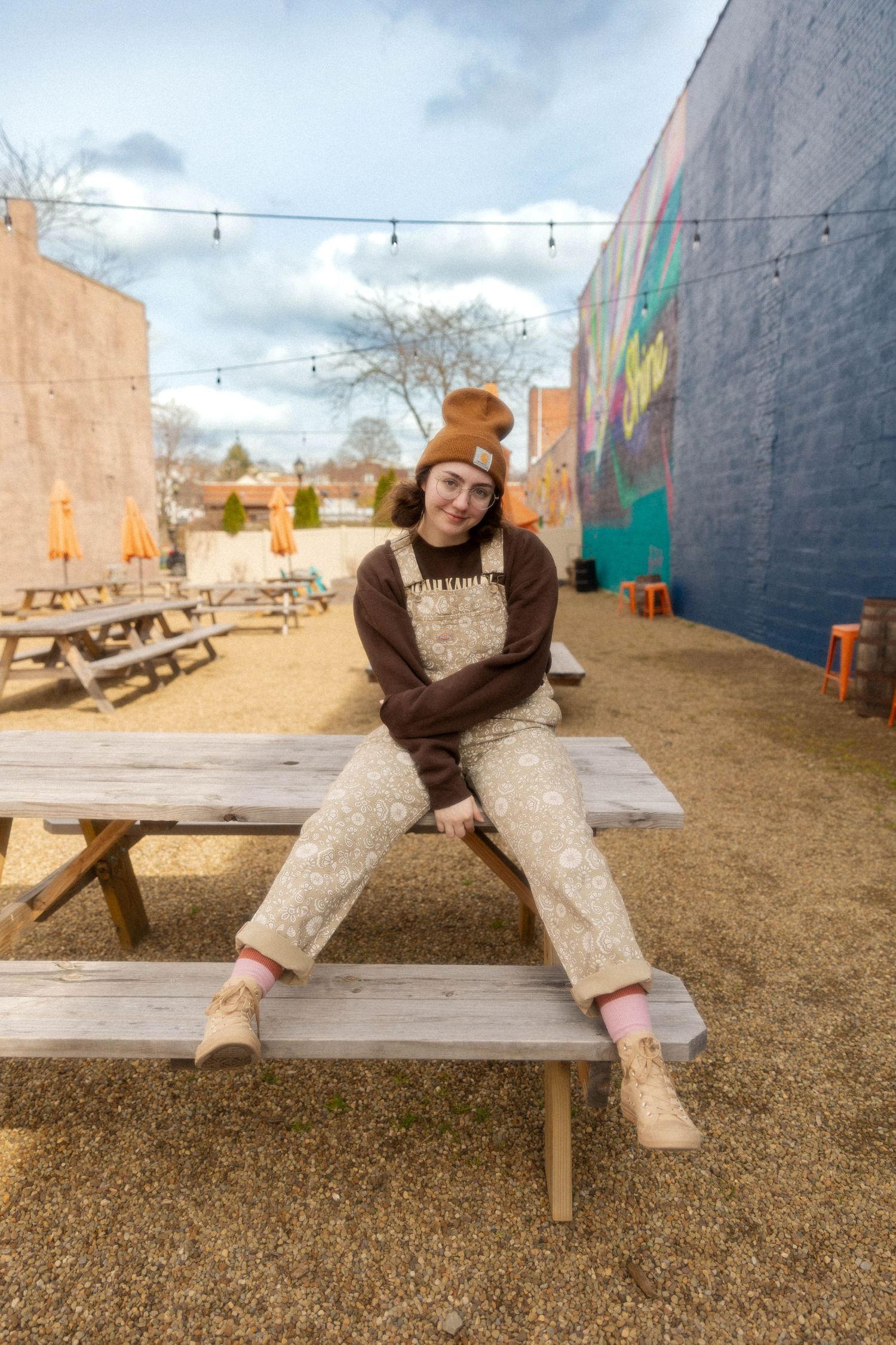 Relaxed pose in brown sweater and patterned overalls on picnic bench with string lights overhead.