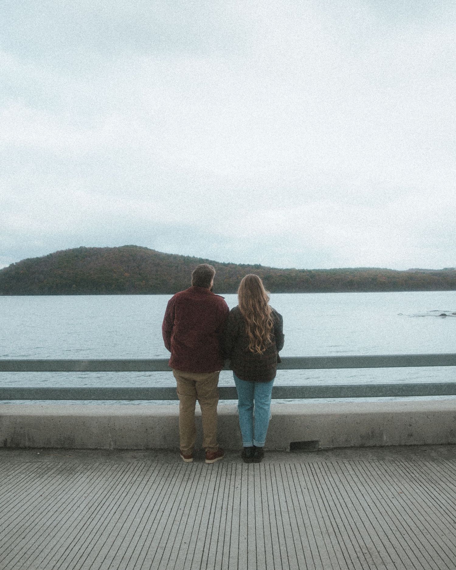 Two people stand at a seaside railing overlooking a misty bay and distant mountains on an overcast day.