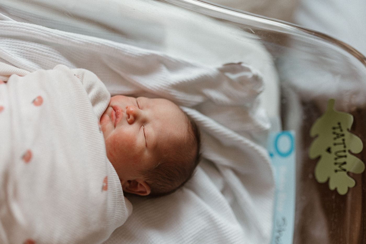 A newborn baby sleeps peacefully in a hospital bassinet wearing a white polka dot swaddle.