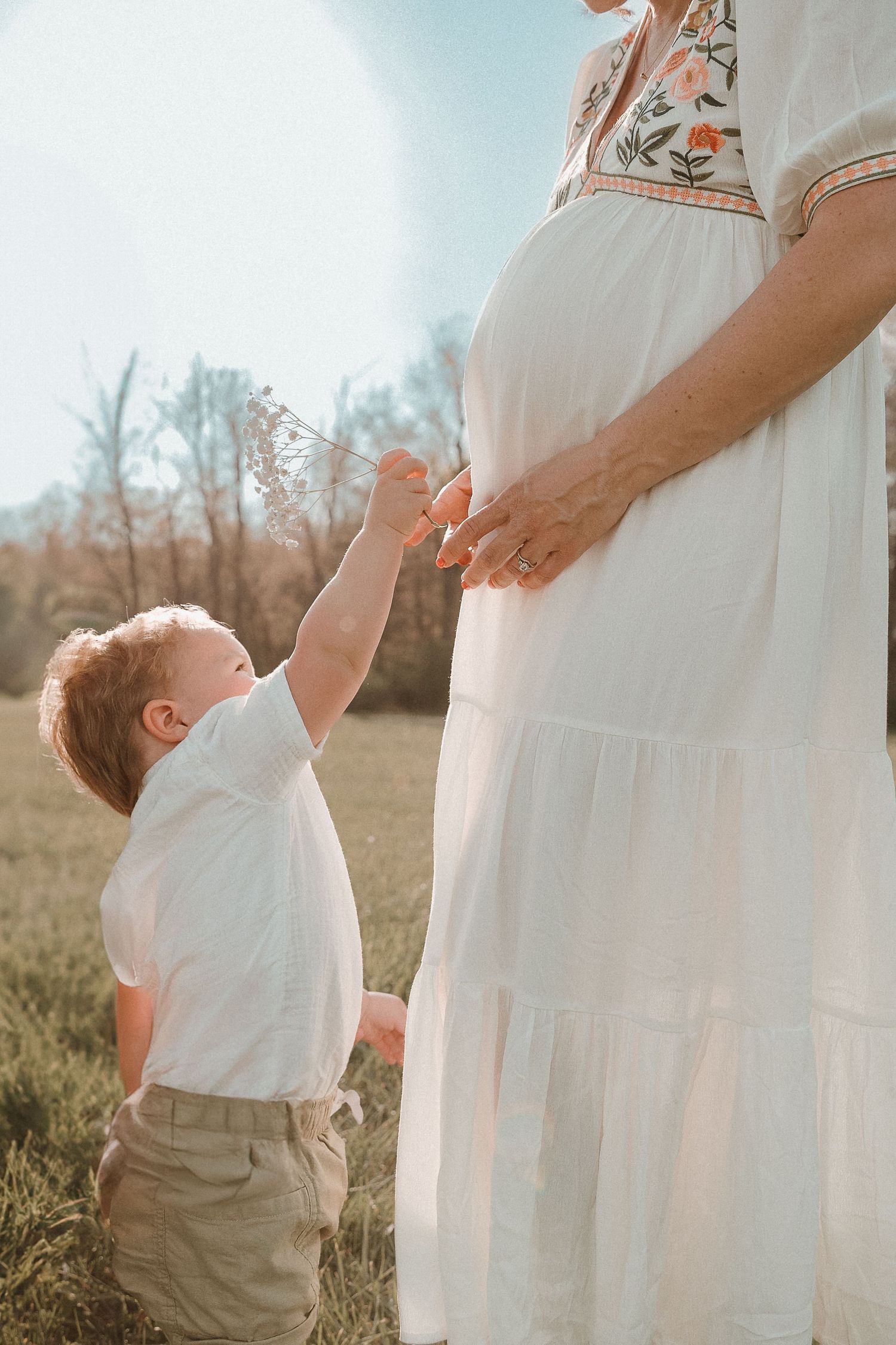 Toddler reaching up to touch pregnant mother's belly in sunlit field,tender maternity session in soft light and earth tones.