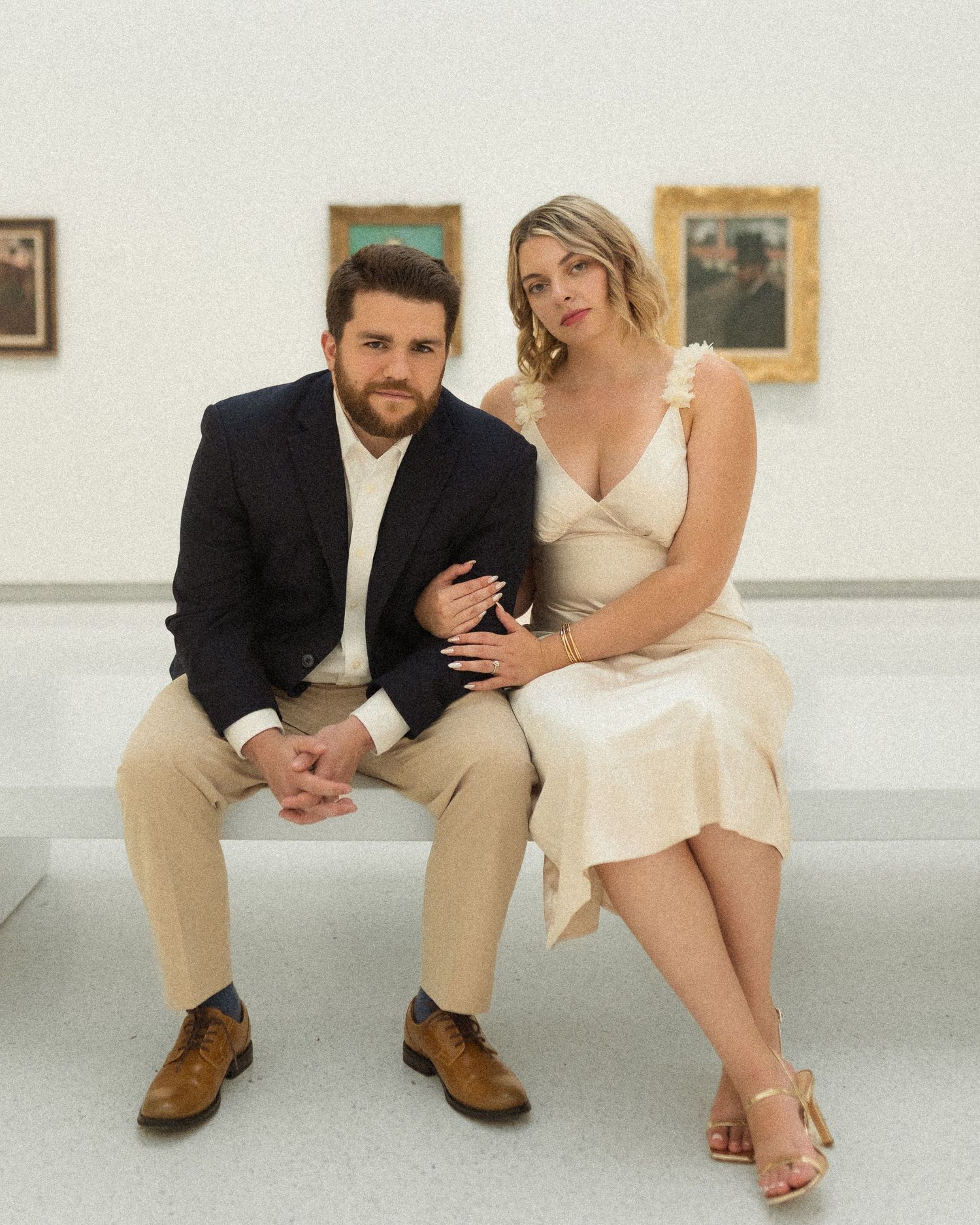 A couple in formal attire sits together on a white bench in an art gallery setting.